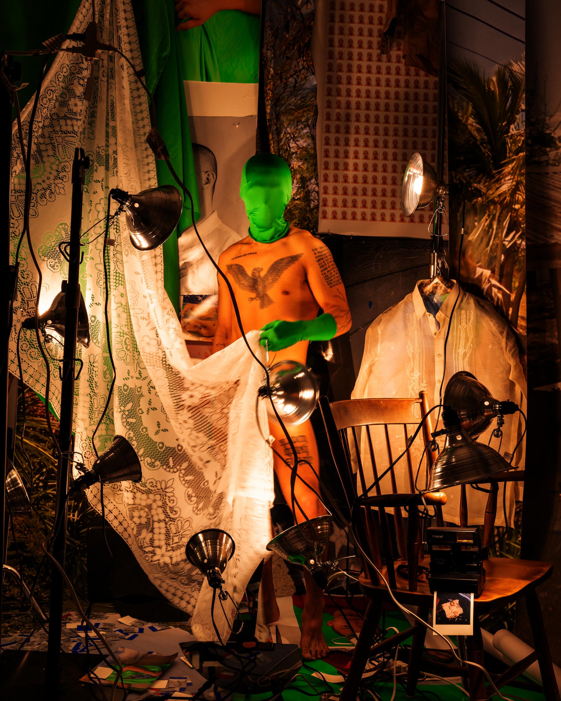 A nude man stands in a crowded room of prints and hot lights. His face is obscured by a green mask.