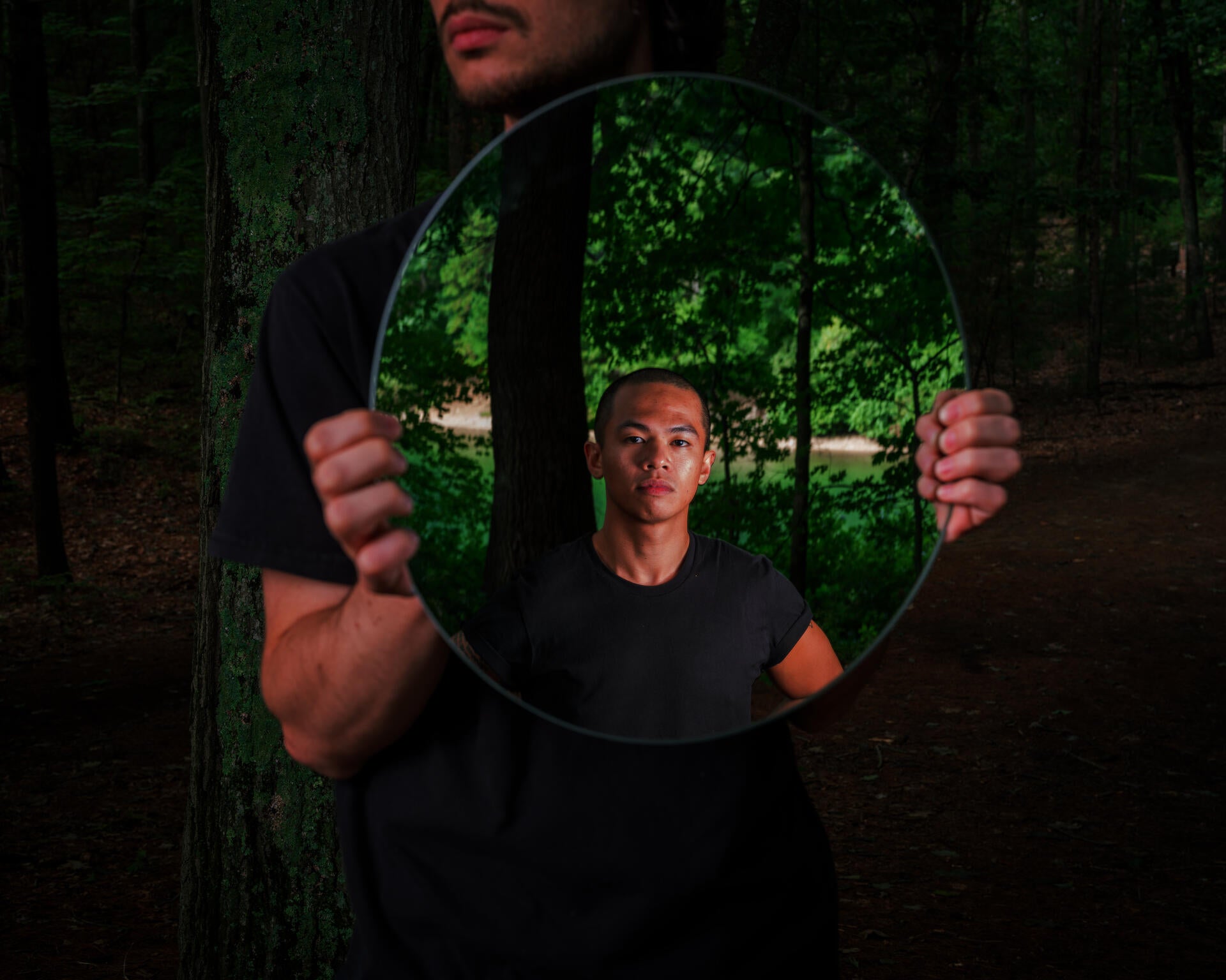 A man in a black shirt holds up a mirror showing another man wearing black. They are in a wooded area.