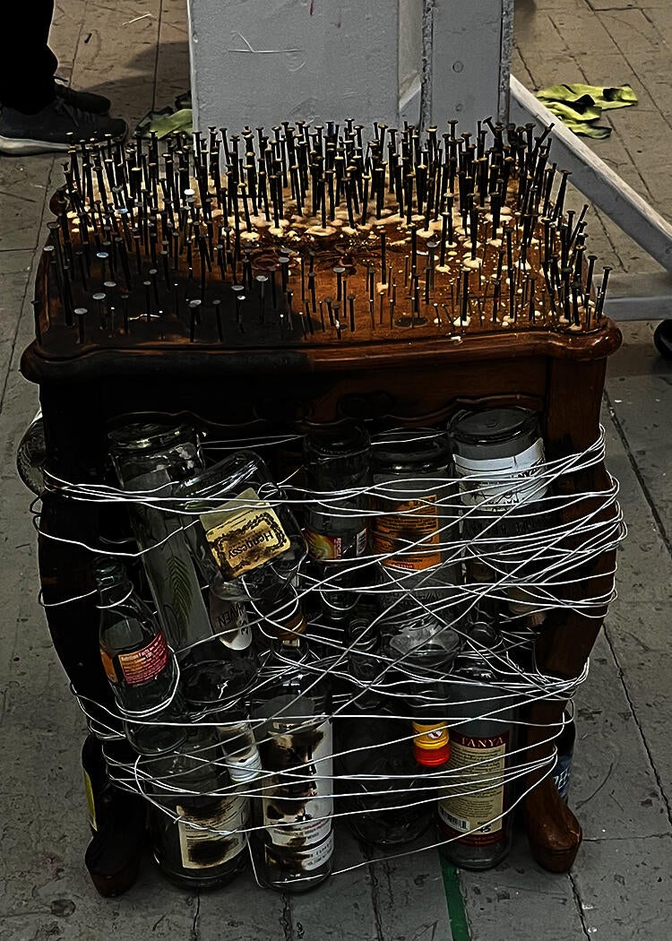 Side table wrapped with with wire and empty alcohol bottles. Hold of the table is burnt and rusted nails cover the top of the table except for a flower printed onto the table