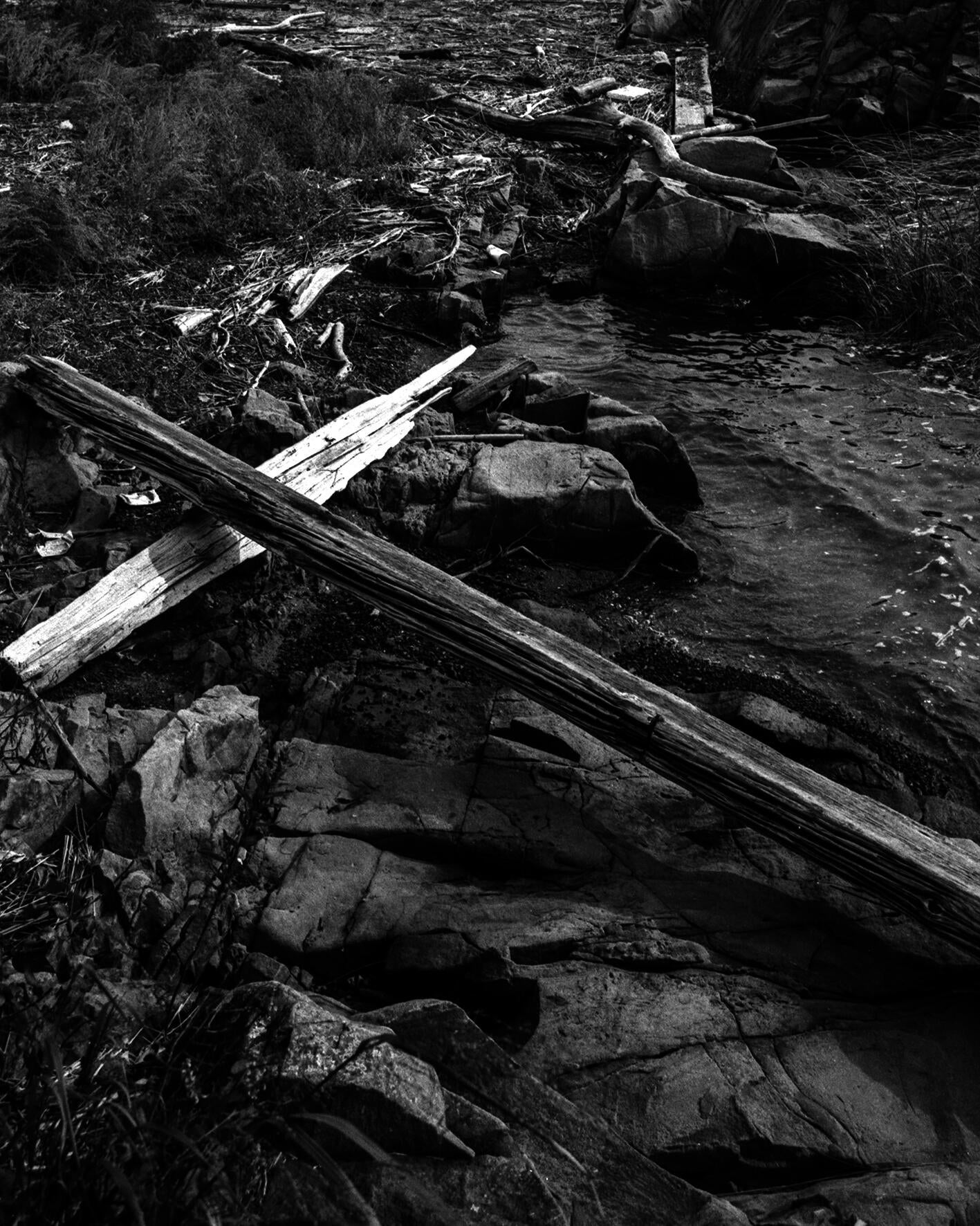 a black and white photograph of two long planks of driftwood in the shape of a cross, washed up on the rocky shores of a bay.
