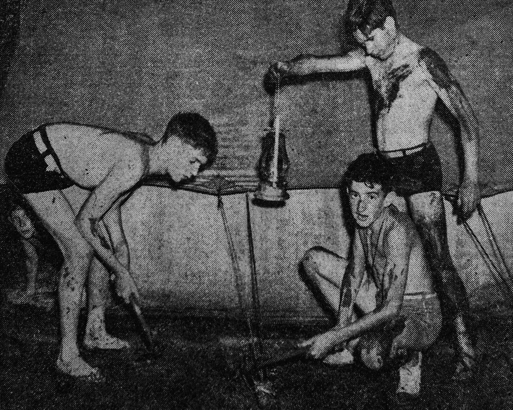 A black and white photo of three light skinned adolescent boys gathered around the side of a tent trying to repair it, it is raining and muddy in the middle of the night.