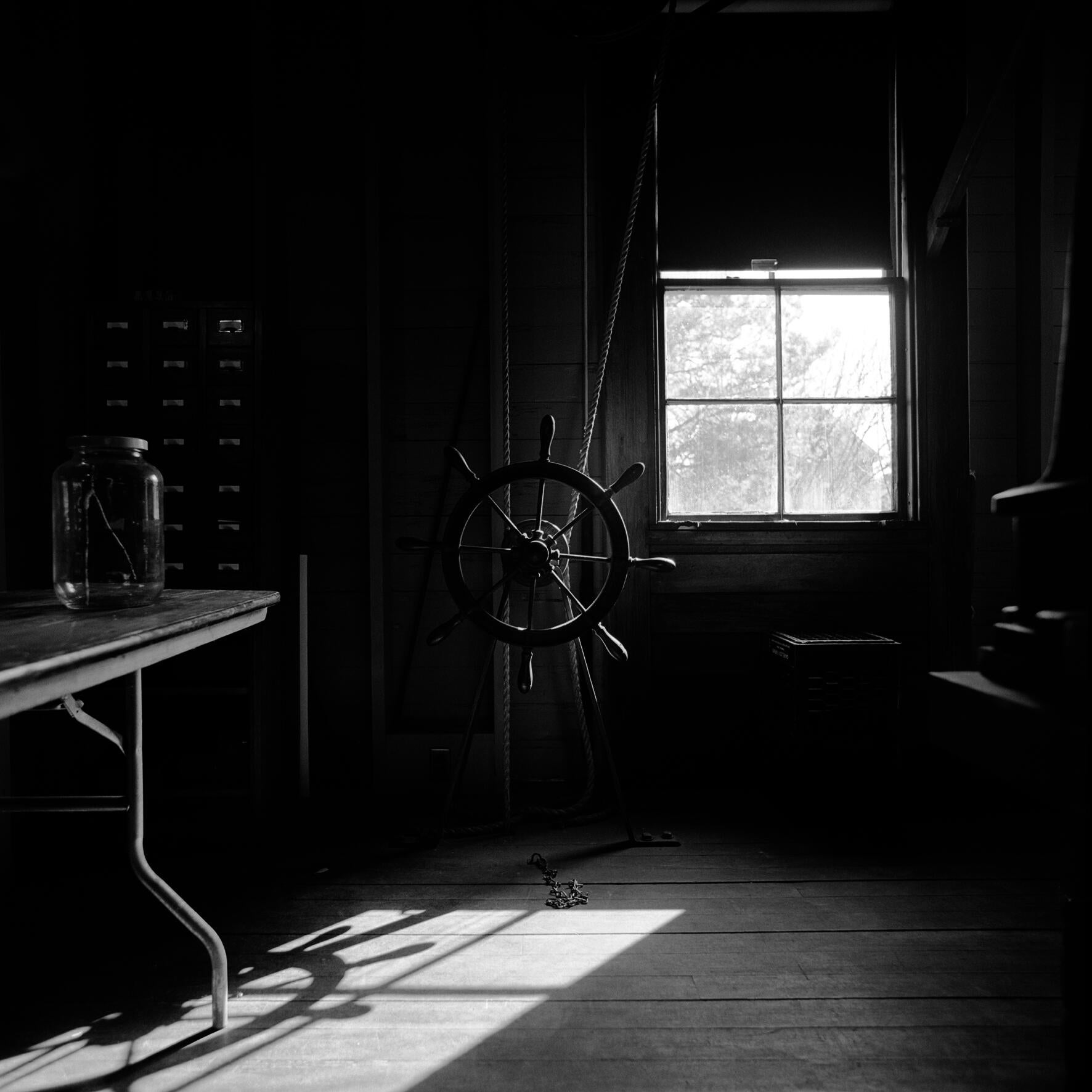 A black and white photograph inside an old looking observatory picturing a wooden ship wheel with ropes attached around it that extend up towards the ceiling.