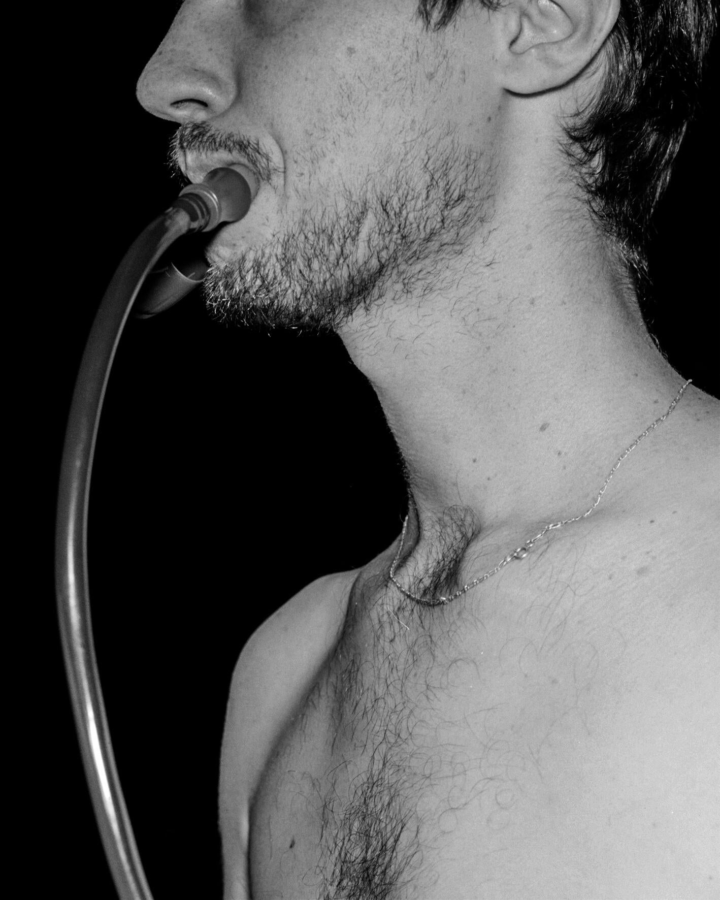 A black and white photograph of the chest, neck and jaw of a light-skinned man drinking from the hose of a hydration flask.