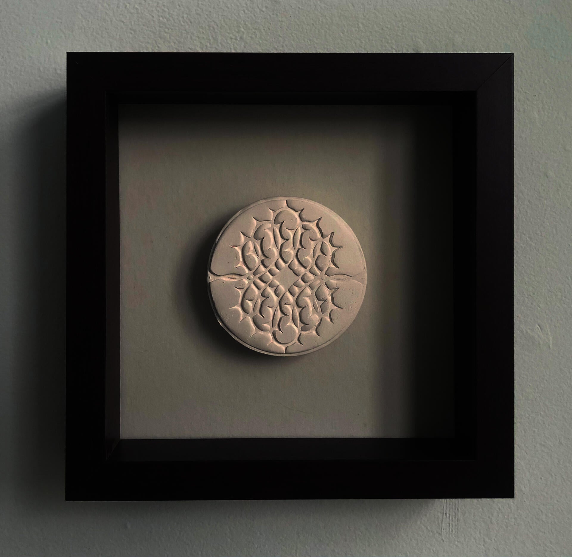 A pale beige ceramic disc with a thorn motif framed in a mahogany shadow box.