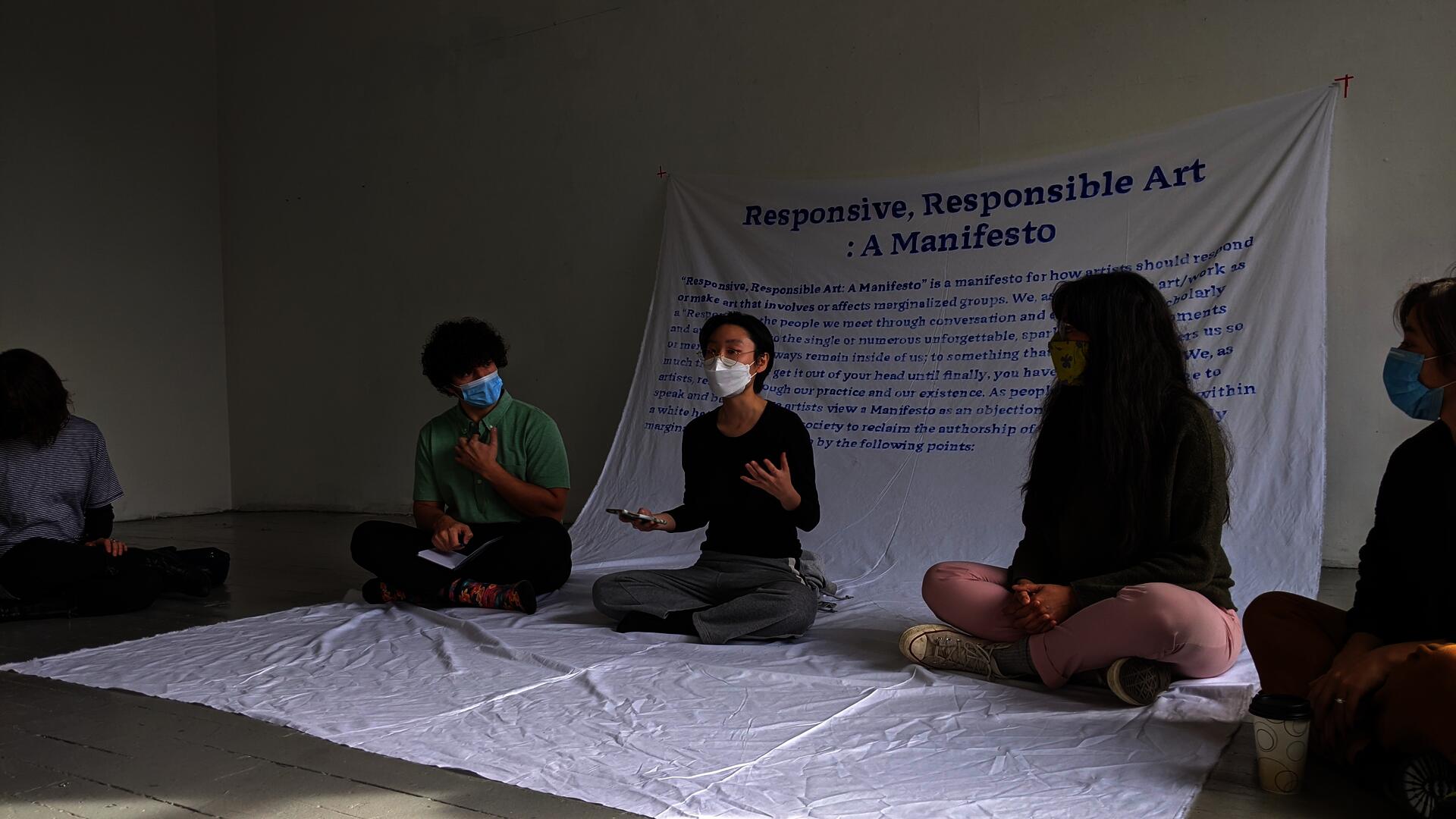Artist reading “Responsive, Responsible Art: A Manifesto” on her phone while seated in a circle with four people on a large white fabric cascading down from the wall, featuring a handwritten paragraph from the manifesto in vivid blue paint.