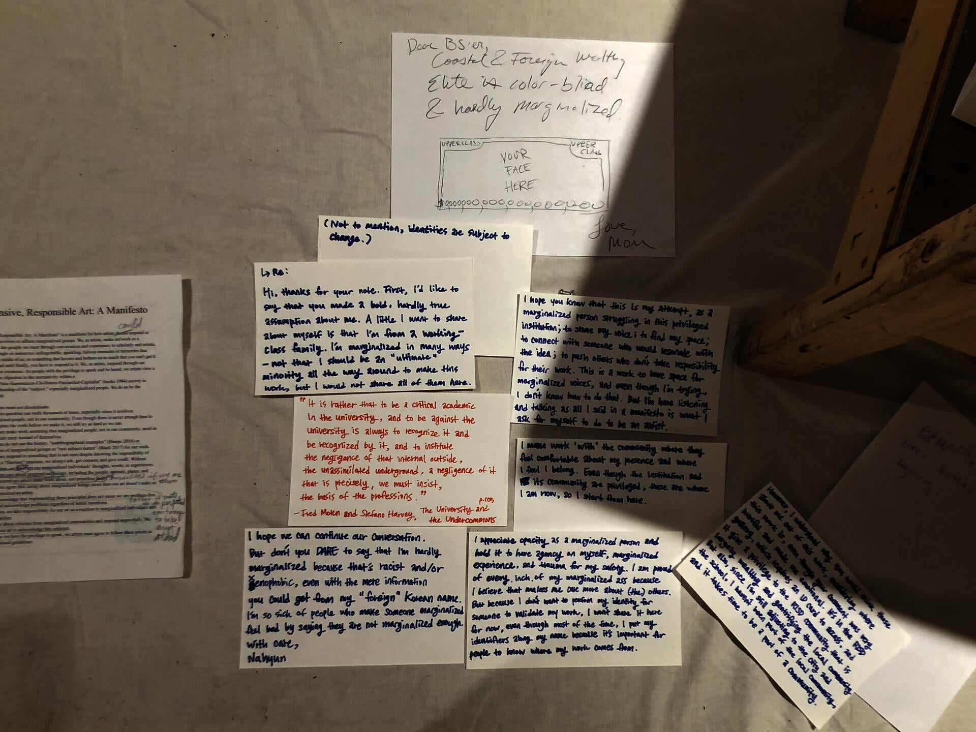 One stack of the printed version of “Responsible, Responsive Art: A Manifesto,” alongside another stack of blank paper, pencils, and colored pencils placed on a rectangular wooden table resting on the fabric banner of the manifesto on the floor.