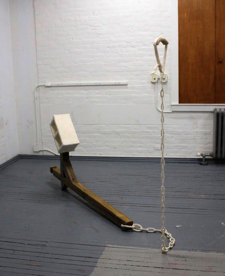 A pine mailbox post lays on the floor, with a felted cinder block balanced on on end and a felted chain and dog collar suspended from the other end.