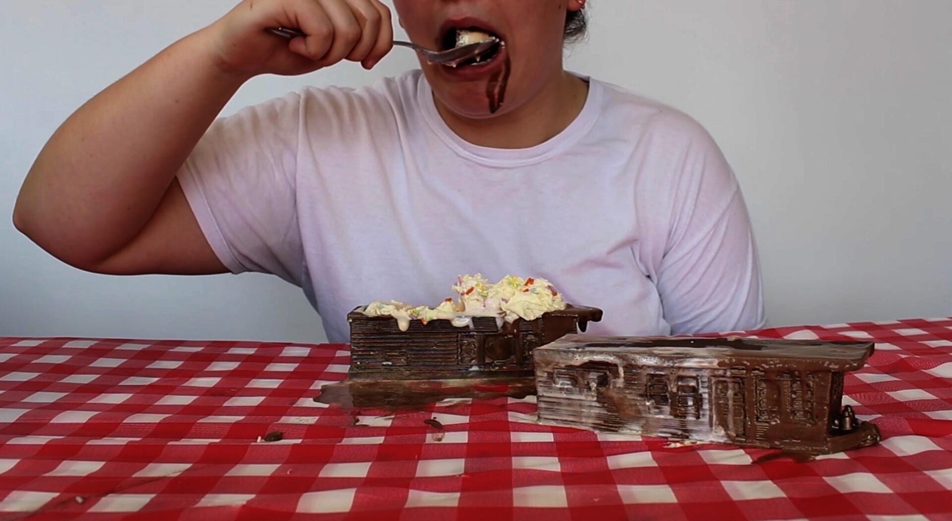 A white person sits at a picnic table behind two miniature bronze trailer homes. The trailers are vessels containing ice cream sundaes. The person inserts a large spoonful of vanilla ice cream in their mouth