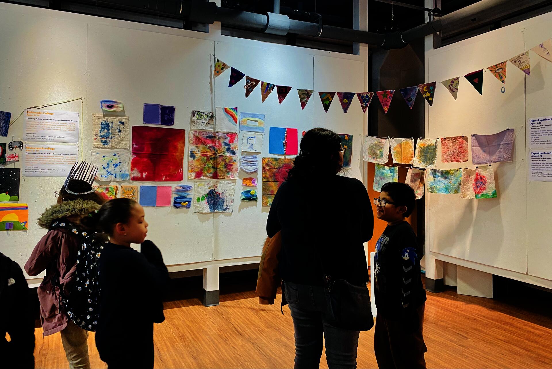 Art show exhibition with student paintings hanging on the walls and students and parents observing the artwork.