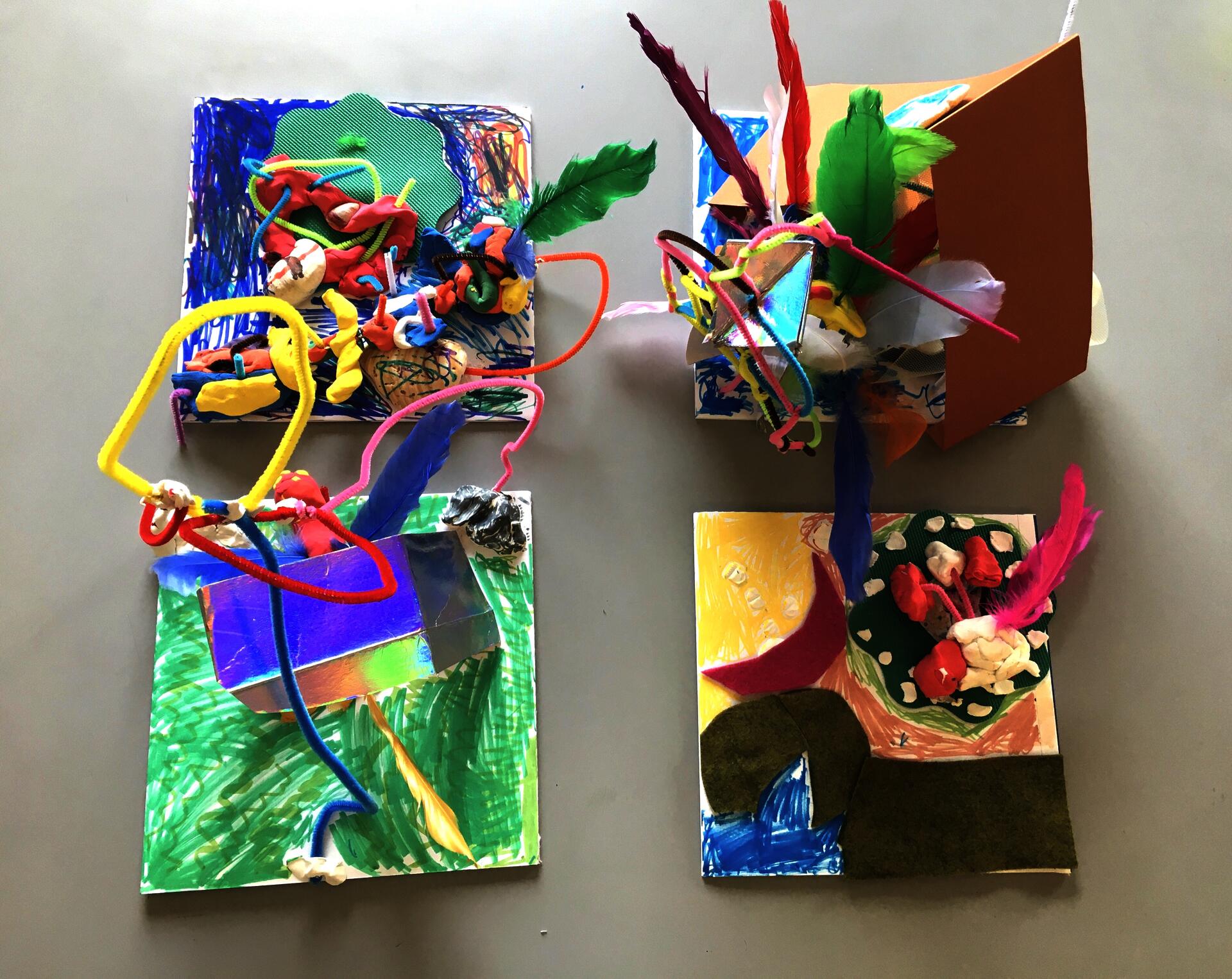 Four "habitats" built on square pieces of foam core, colorful air dry clay, pipe cleaners, shells, feathers, repurposed fabrics and paper, marker. Each habitat is constructed and colored in its own way, with some elements extending upwards towards the camera.