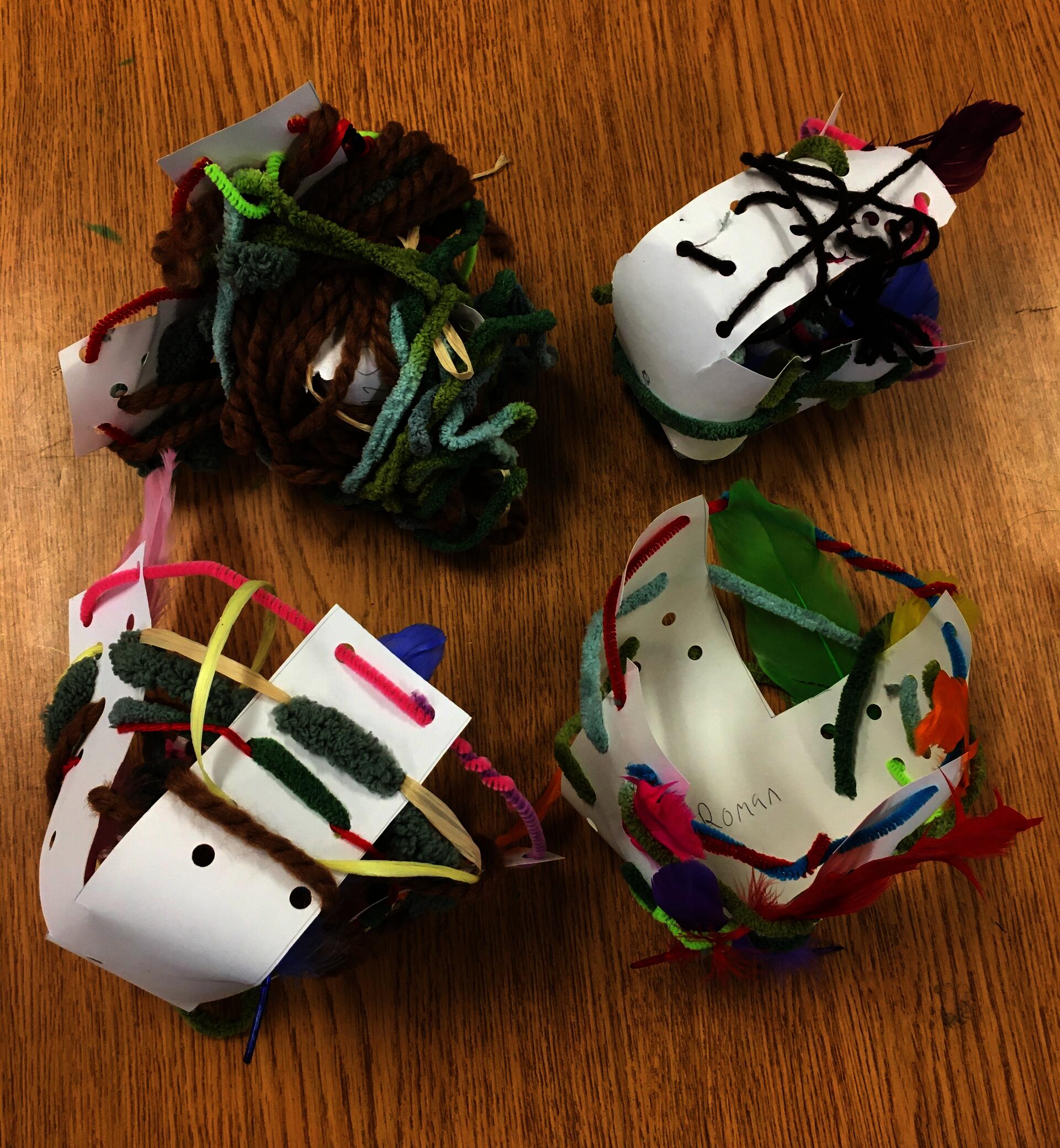 Four nests made of pipe cleaners, various yarns, raffia ribbon, and feathers woven through and around cupped x-shaped frames made of hole-punched white paper. Each nest has its own style and color scheme.