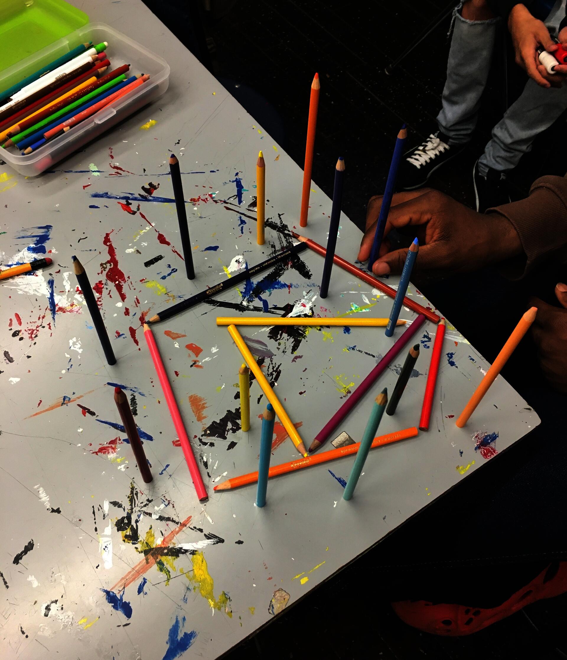 A student’s hand placing one of 13 colored pencils that are standing on their ends, pointing vertically upwards from a paint-splotched table. The vertical pencils surround 3 colored pencils lying flat on the table in the shape of a triangle, inside a pentagon made of 5 colored pencils. Another student’s legs and hand can be seen in the corner of the image.
