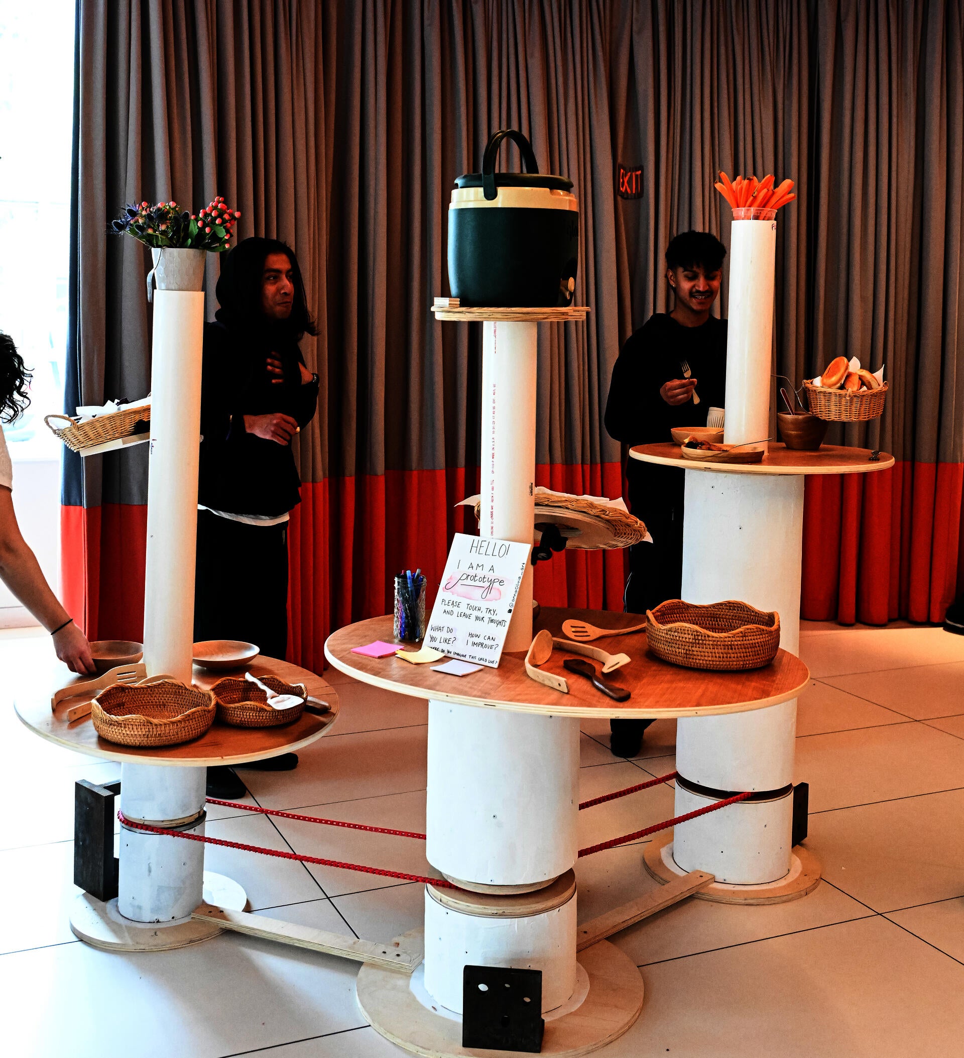 Three white columns with ovular tabletops and one tall cylindrical pipe coming out from each surface. The three tables are connected by a red mechanical belt which turns them. 