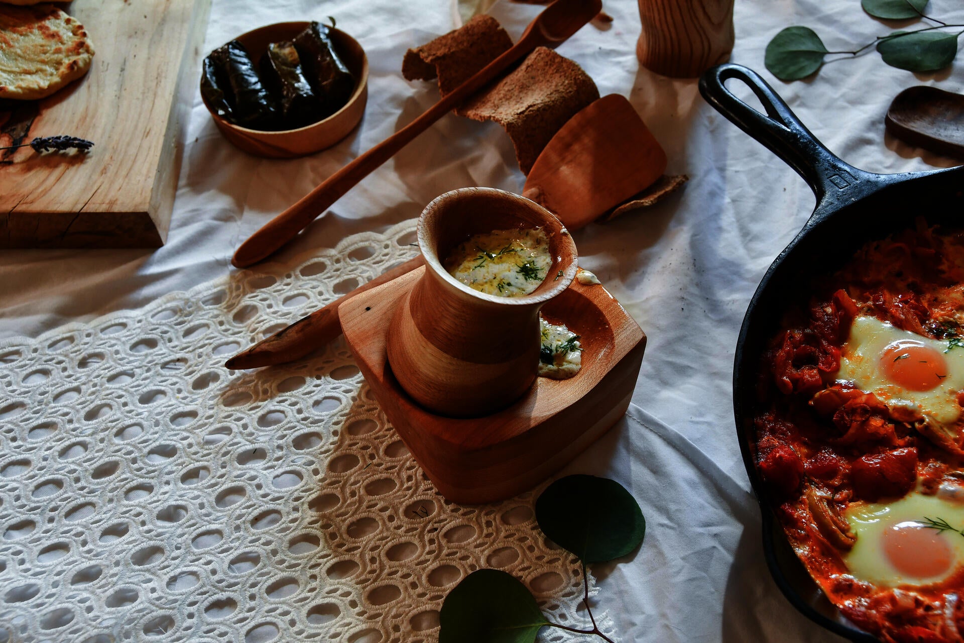 Wooden goblet filled with tzatziki and set into a larger wooden square bowl with two indents, wooden spoons, and part of a larger table spread on a white lace tablecloth. 