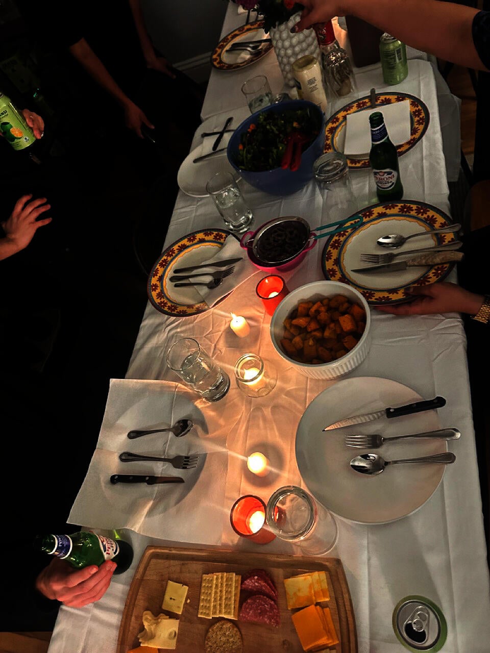 A long rectangular dinner table set with a white tablecloth, large round plates, and cutlery laid out in various patterns and orientations. 
