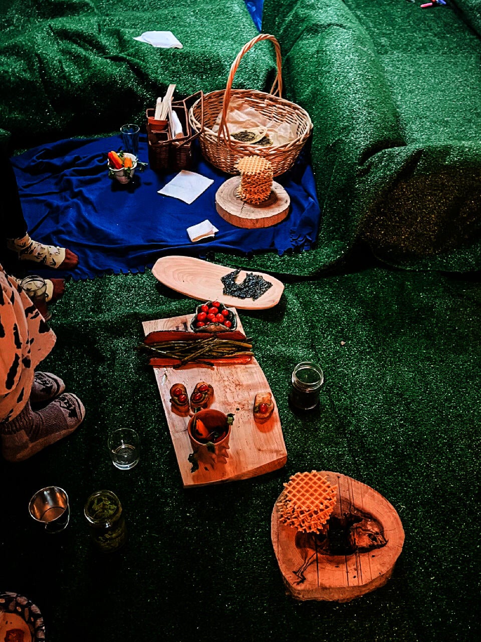 The green astroturf-covered floor of the apartment used for the indoor picnic. A blue blanket with a basket and a stack of waffle cookies, and other wooden food platters.