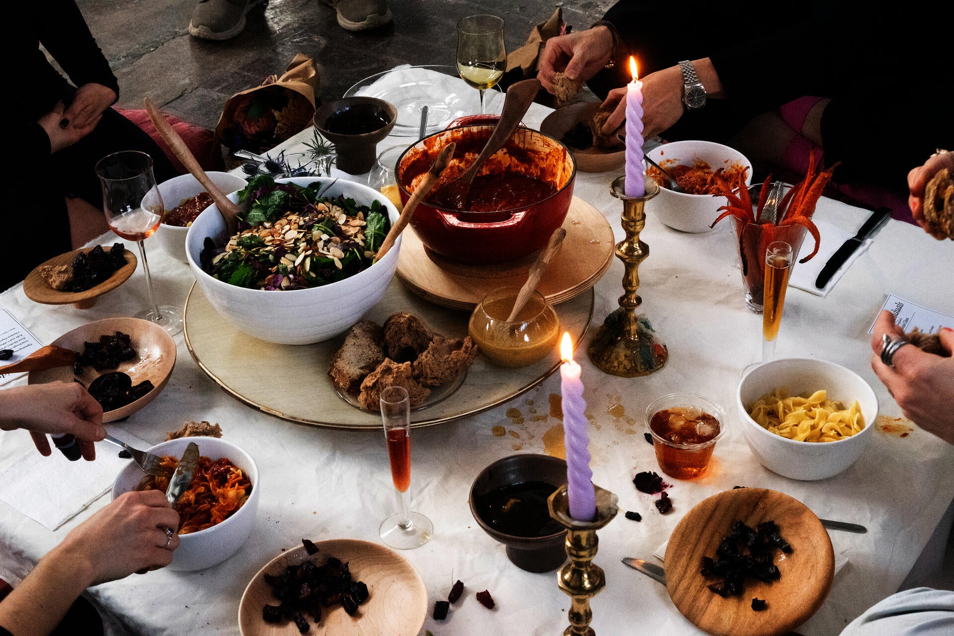 A close up detail photo of a low table with four people sitting around having a very colorful meal of salad, beets, chicken paprikash, purple candles, and with a wooden turntable in the middle. 