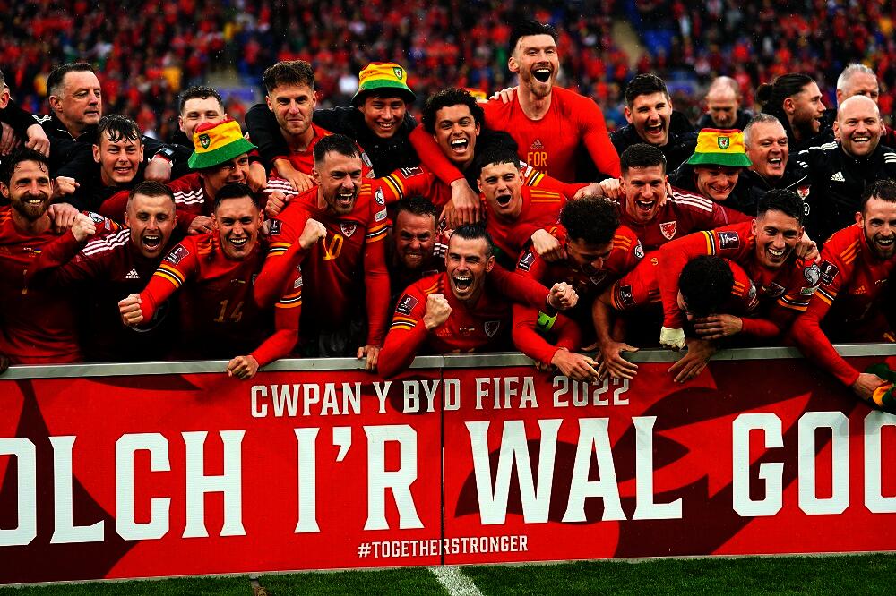 Welsh football players wearing bucket hats and celebrating in front of a banner with the words "thank you to the Red Wall" in Welsh.