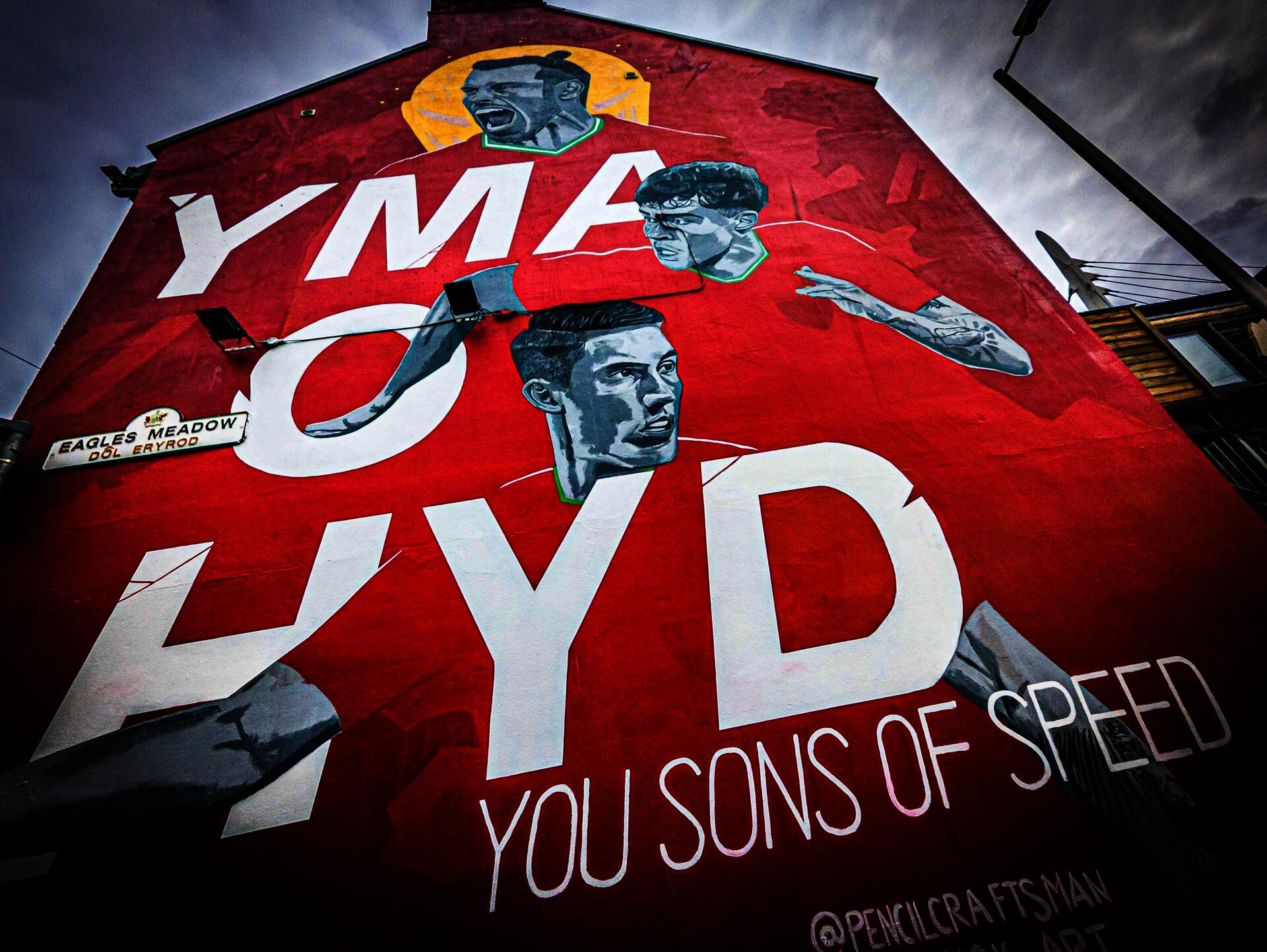 Mural of Welsh football players Gareth Bale, Neco Williams, and Harry Wilson on the side of a building with the words "Yma o Hyd You Sons of Speed."