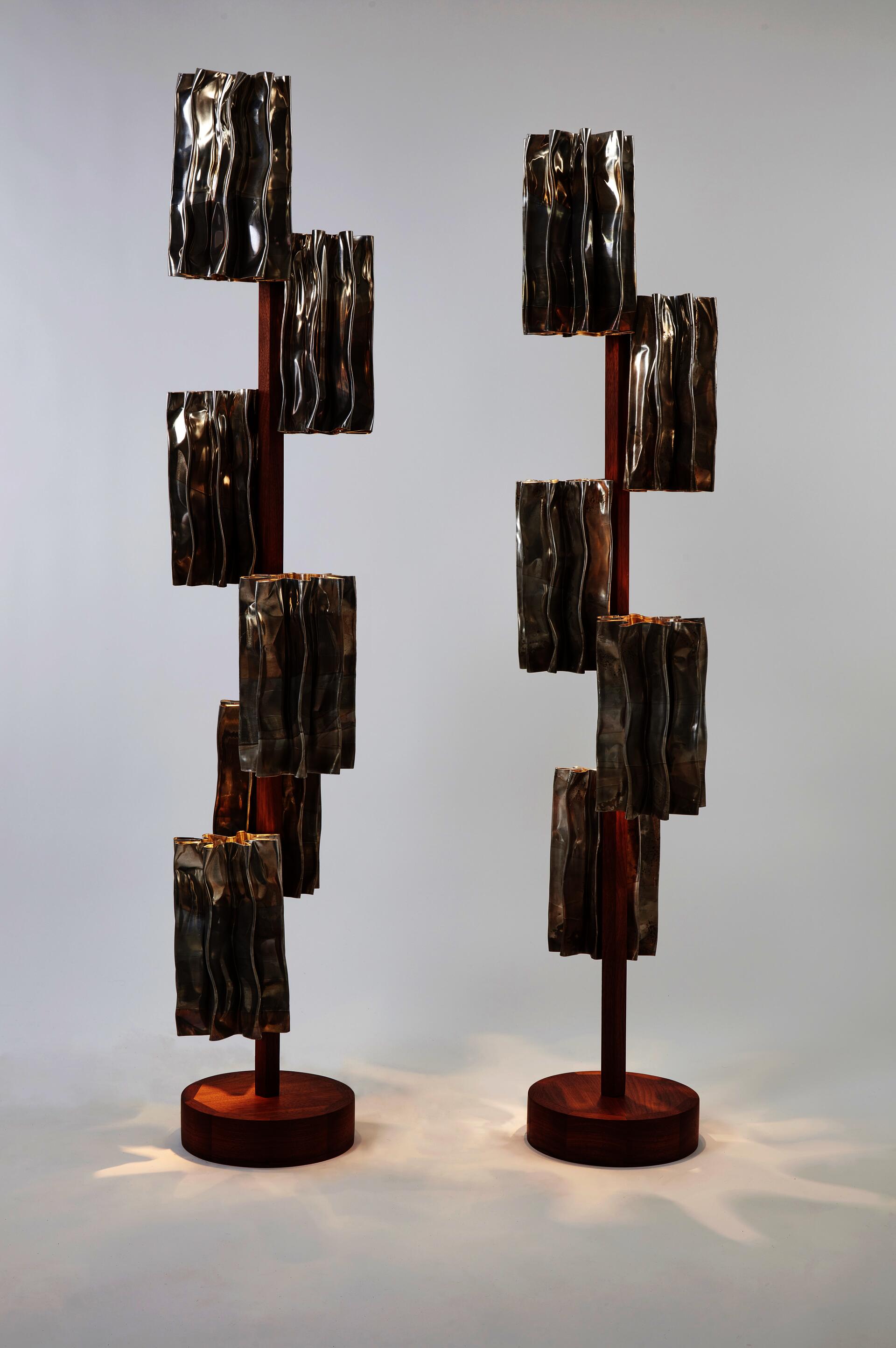 Two modern floor lamps display similarly styled metallic aluminum segments as decorative shades with a central walnut pole and base.