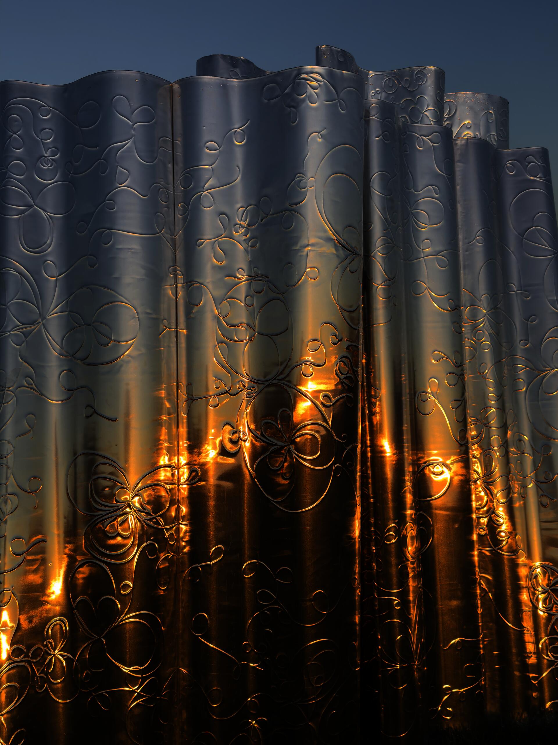 Detail of a metallic, wavy privacy screen with intricate floral patterns reflecting the golden hues of a sunrise. The surface creates a shimmering effect, capturing the light and adding texture to the smooth, undulating forms.