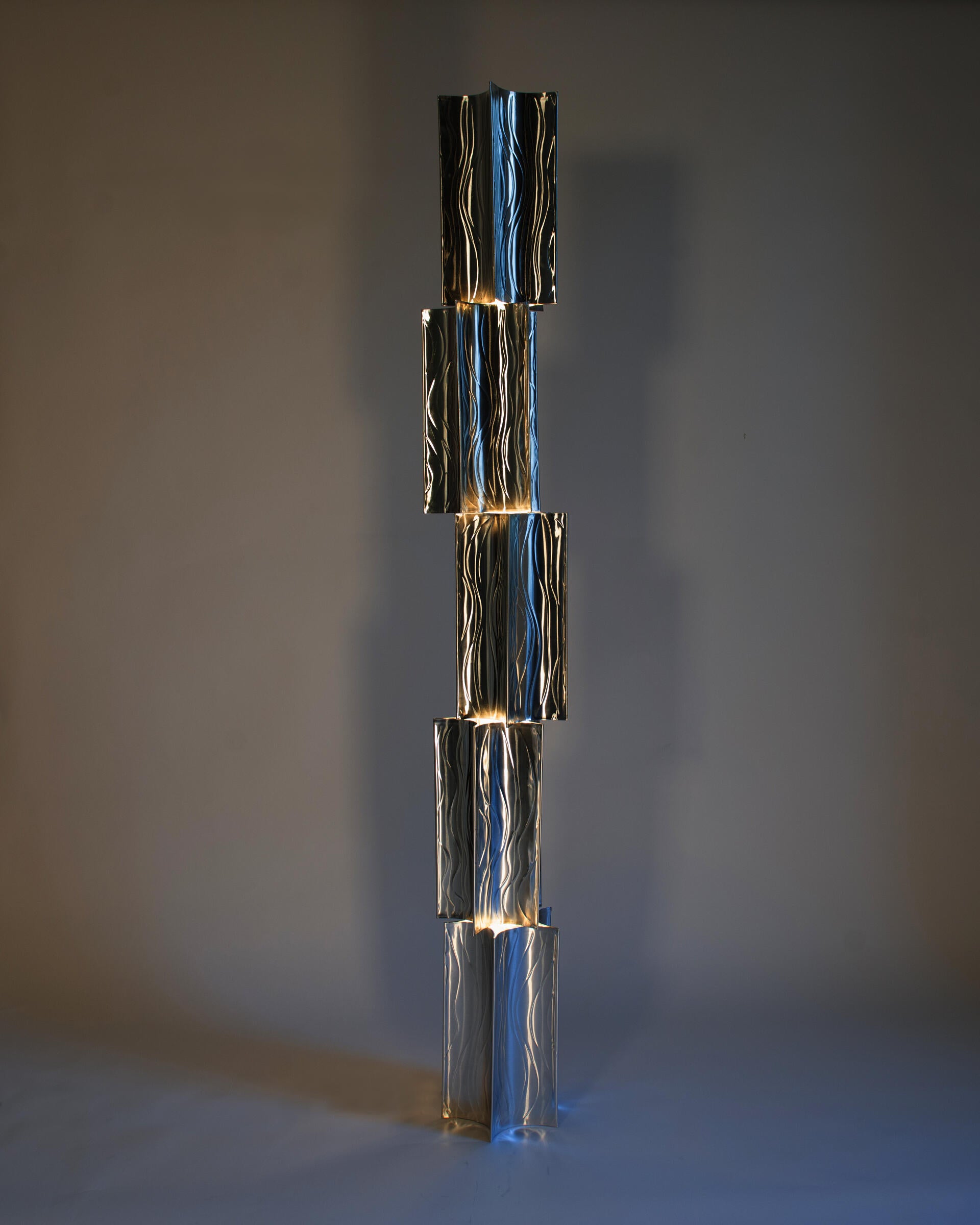 A sleek, modern floor lamp composed of a vertical stack of five aluminum lanterns. Each panel has a wavy texture and emits a subtle, warm light from the edges, creating a cascading effect.