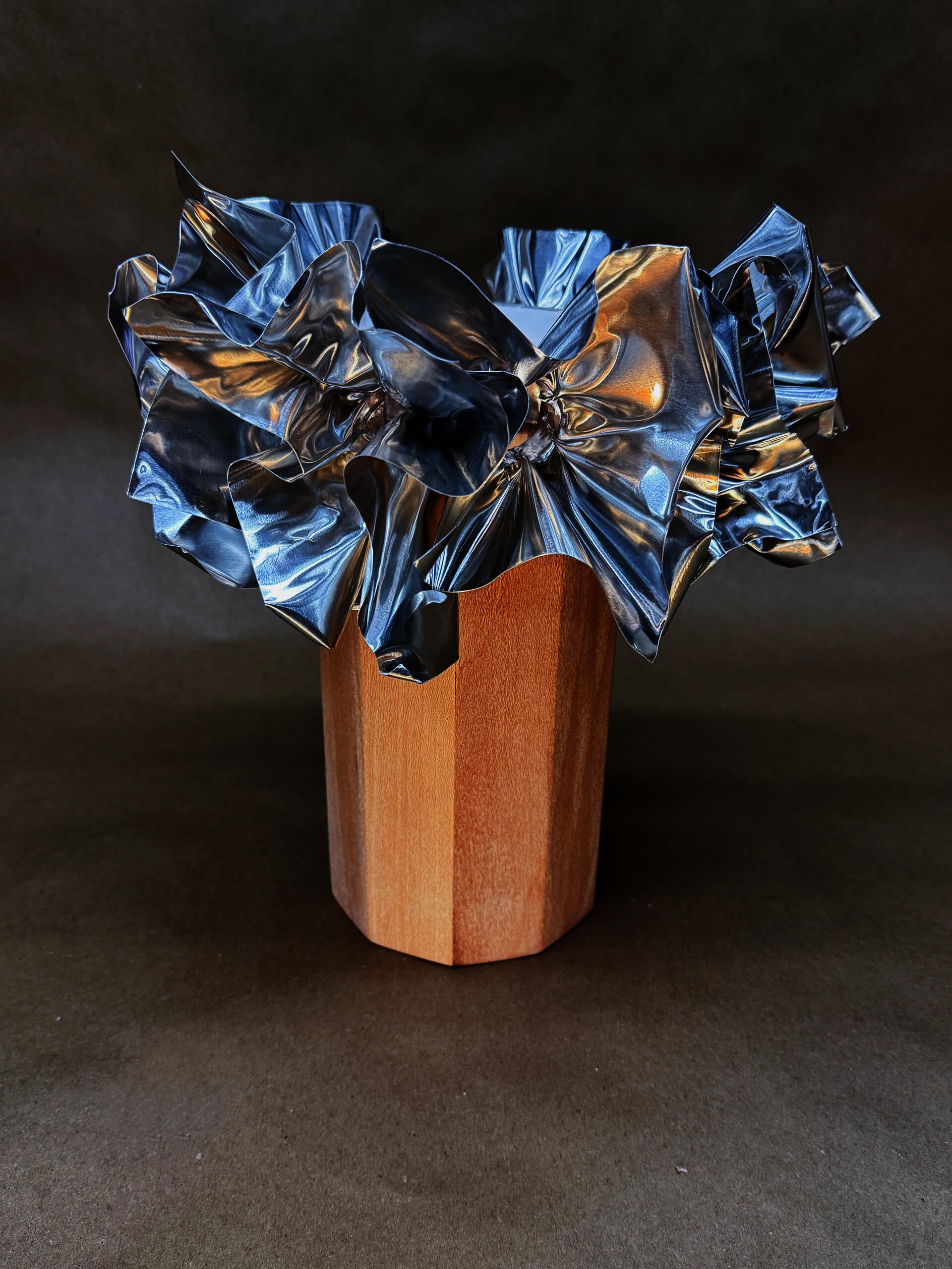 A geometric wooden lamp base contrasts with the lampshade, intricately crumpled and shiny aluminum sheets resembling abstract flowers