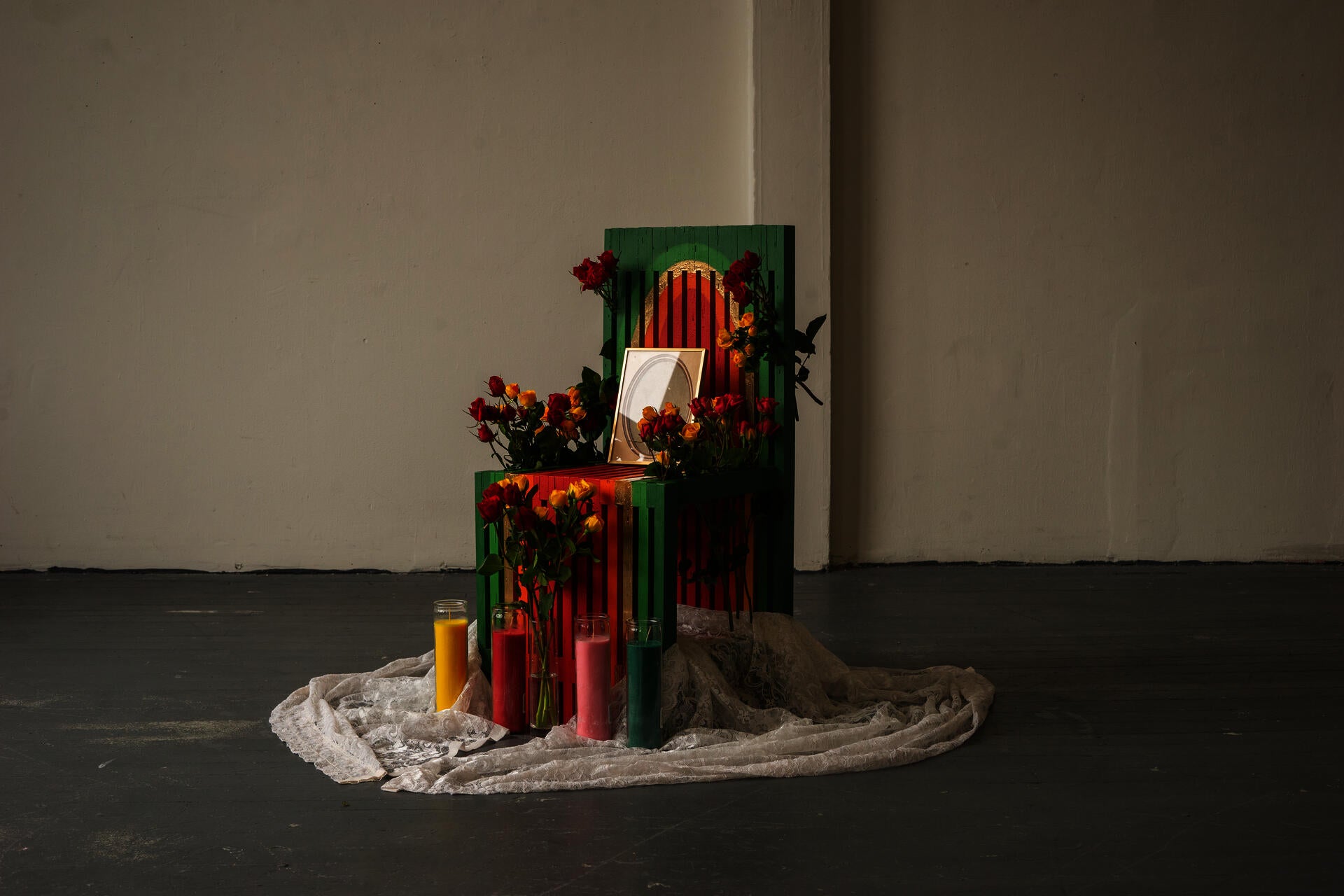 A slatted chair painted in a warm gradient sits in the middle of a stark room. Flowers are weaved between the slats, with a doilie, prayer candles, and flower vases sitting at its legs.