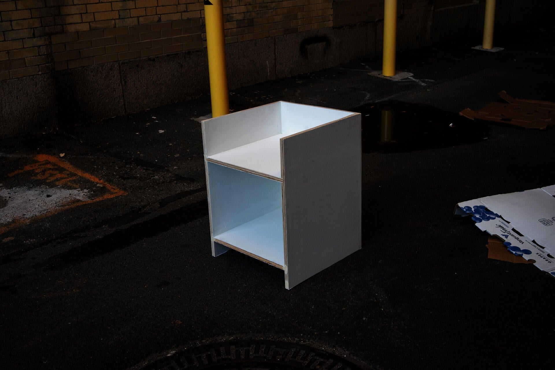 A stark white chair in a rigid geomgetric form sits in a dark alleyway.