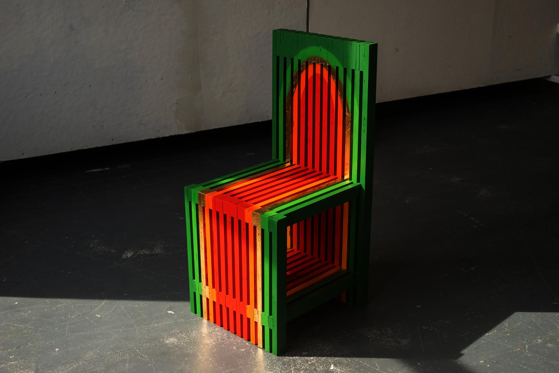 A colorful slatted chair painted in a colorful warm gradient sits in the middle of a stark room.