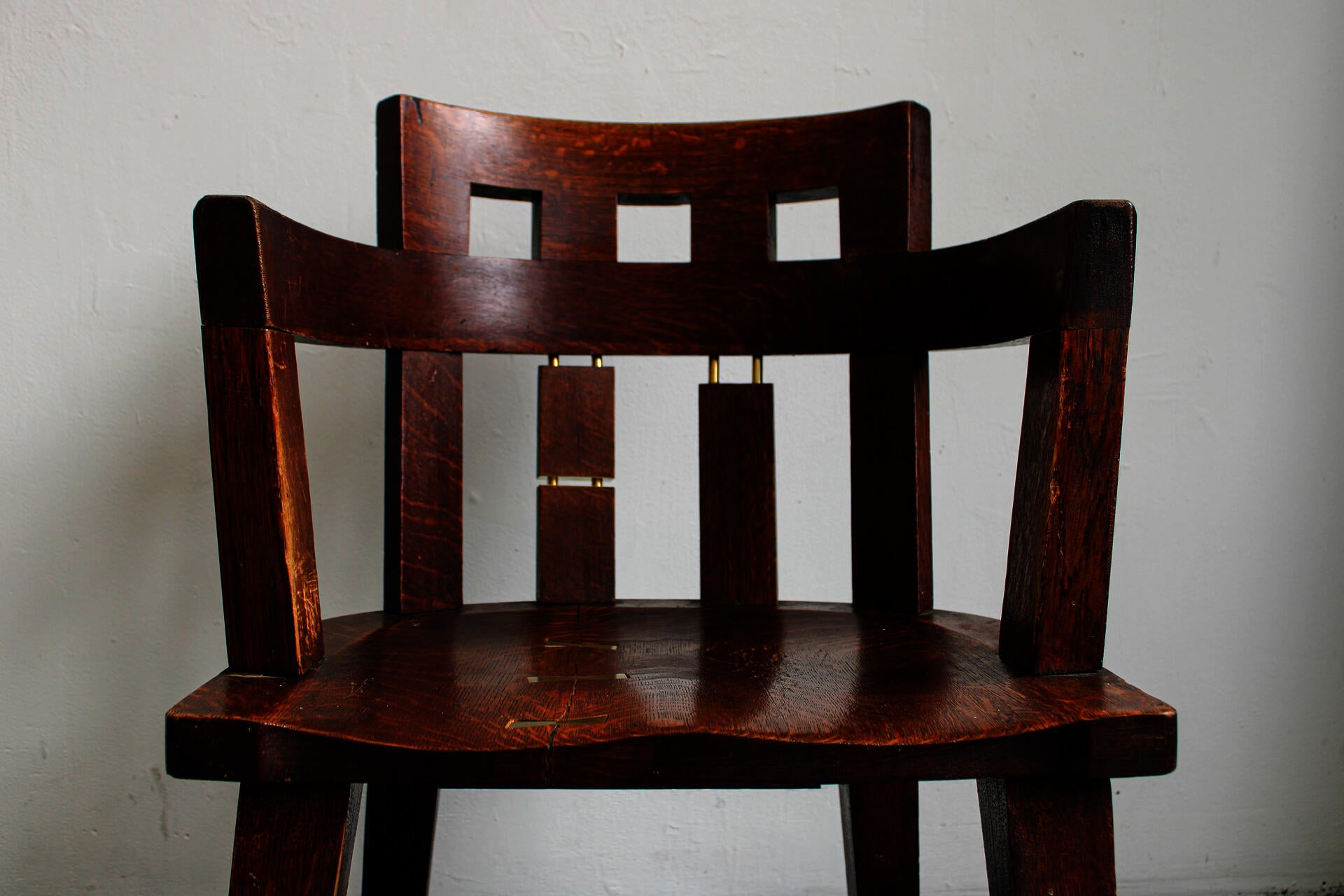 A weathered, dark brown oak chair is repaired with shiny brass metal. Brass bowties are embedded into seat where it cracked. Brass dowels show through several of the chair's legs and back dowels.