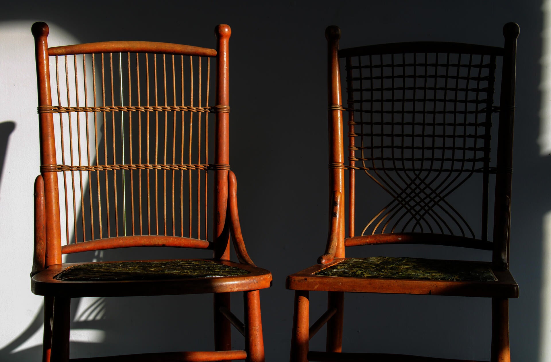 Two chairs (dark brown, ratan-style) sit side by side. Both have repaired seats made from woven pieces of kelp. The kelp reads various hues of green, from dark to light. Somewhat like a camoflauge pattern.