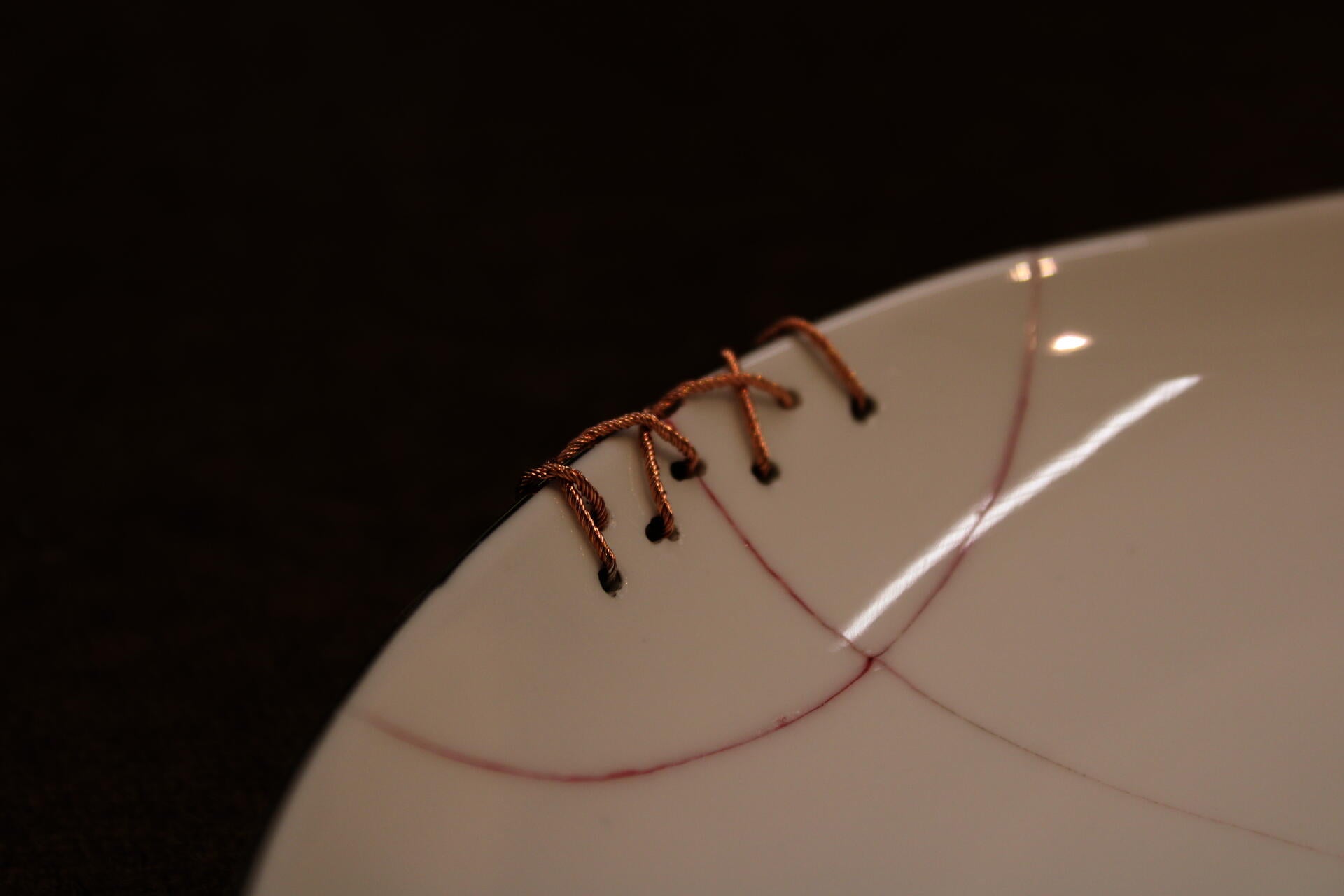 Close up image of the edge of a white plate which has been essentially sewn back together using tiny copper wires. The crack on the white plate is highlighted with a subtle red epoxy filling.