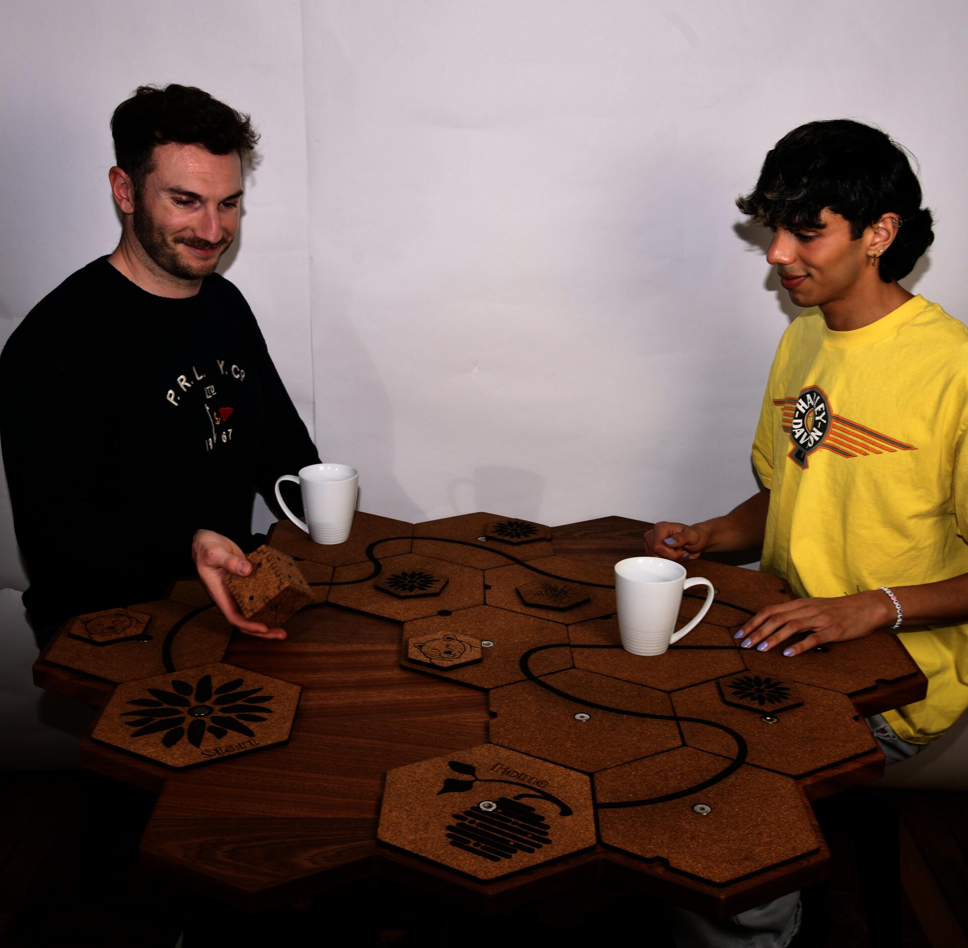 Two people, playing a game on the table, sapele table, honeycomb table, sitting together, one person moving cork tile, a white mug sitting on the table top