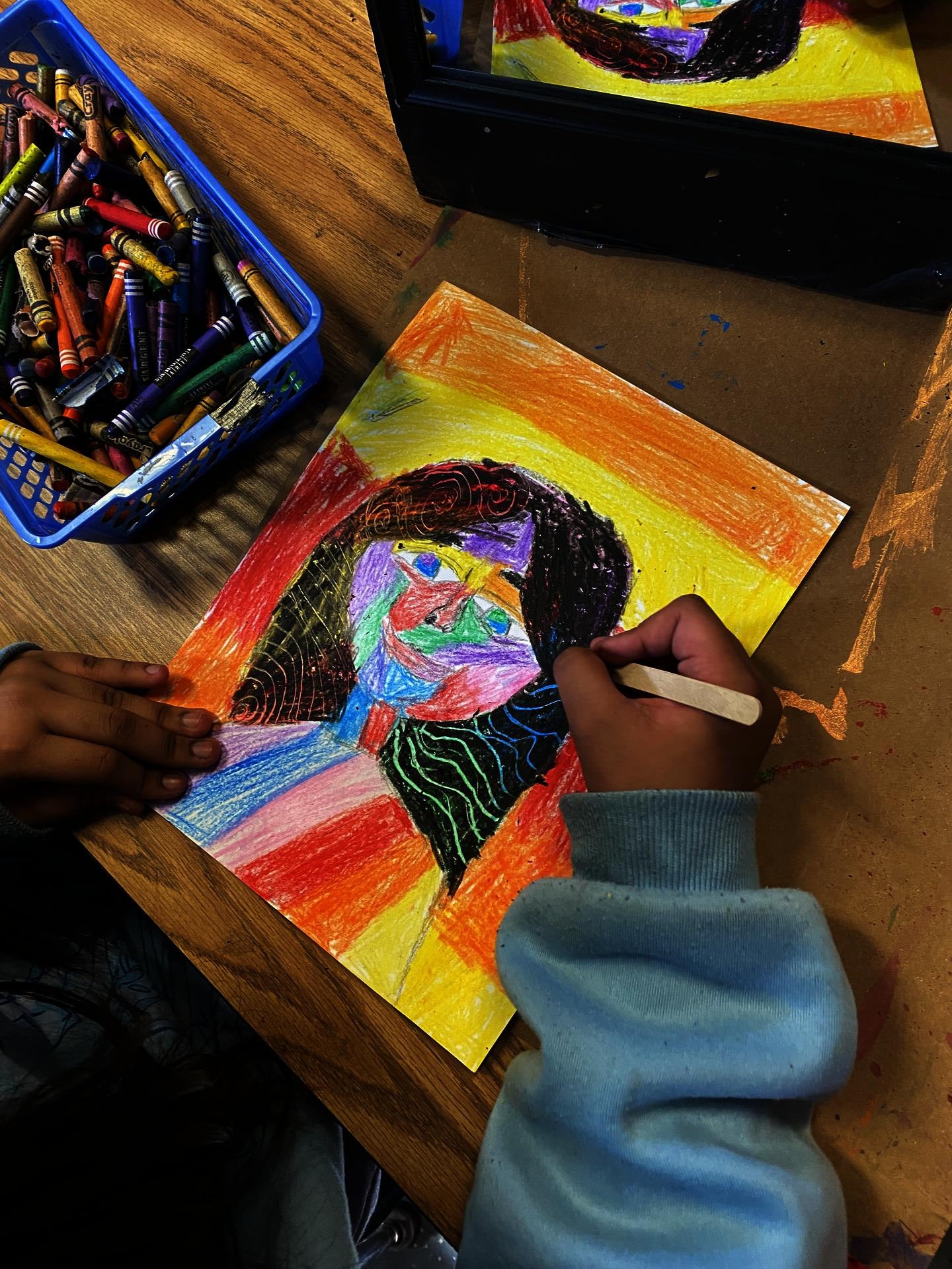 A student's hand holding a popsicles stick is scratching into a crayon-laden paper. The popsicle stick is creating patterns in the self portrait. A bin of crayons can be seen to the left of the image.