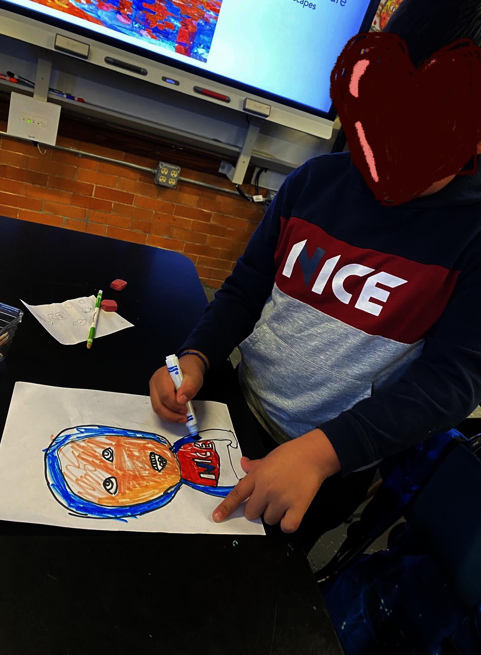A student with a heart over their face to protect privacy is standing at a table and coloring with marker a portrait of themselves. The student is using blue, red, and a tan colored marker.