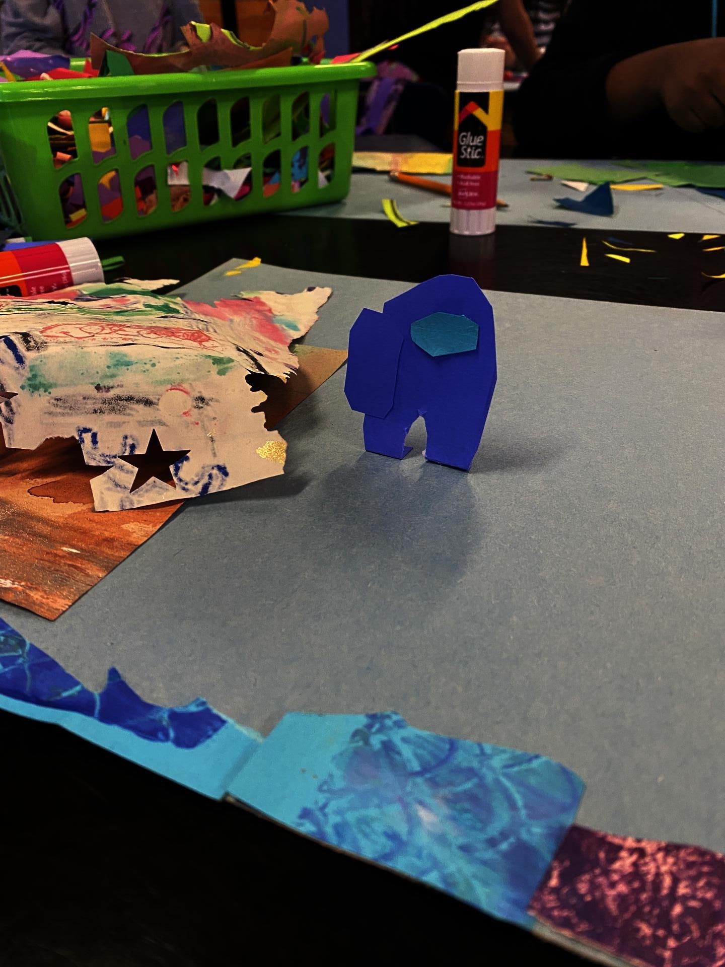 A paper collage cut out of a spaceman from the video game Among Us stands up in a 3D fashion on a student's paper. Seen in the background is a mess of papers and gluesticks.