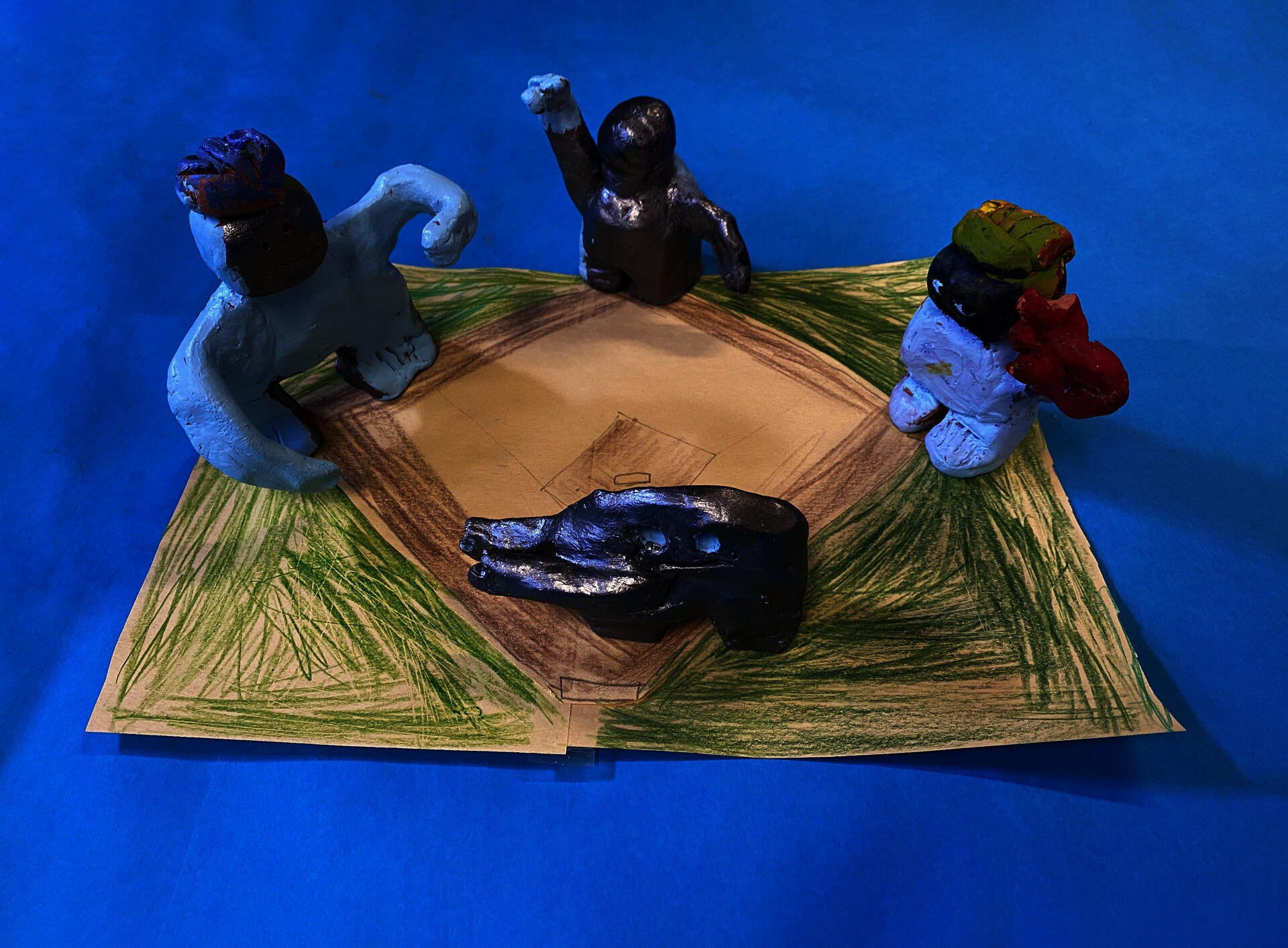 Four clay figurines stand on a hand drawn baseball diamond atop a bright blue backdrop. The figurines have been painted a combonation of silver, light blue, and red, and all have some version of outstretched arms.