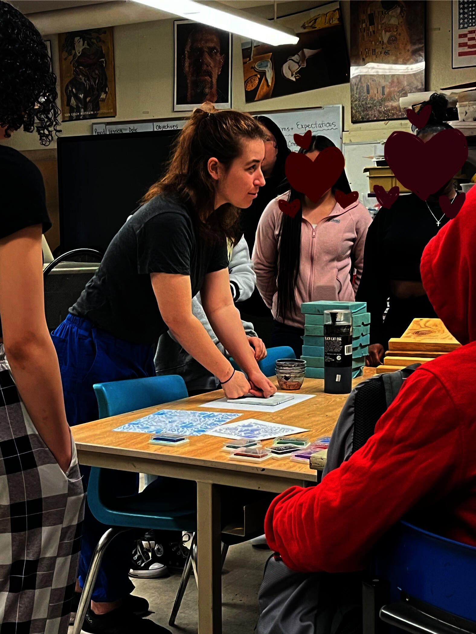 A woman in the center of the image is surrounded by a group of high-schoolers. The woman is pushing down on a sandwich of thin easy-cut linoleum block and a piece of paper onto the table.
