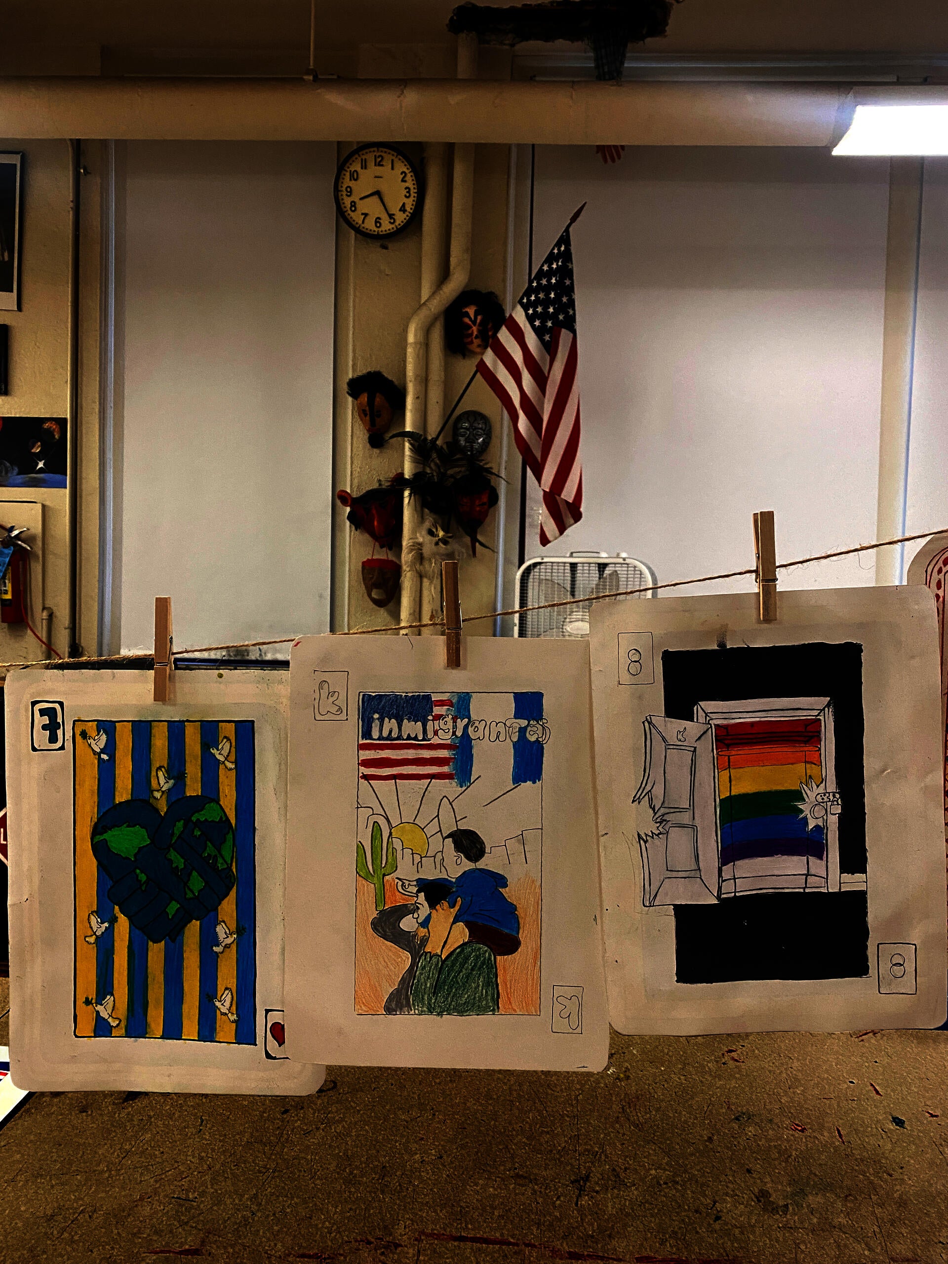 An assortment of drawings in the format of playing cards are suspended in from on a classroom wall with an American flag. Topics of the card include world peace, pro-immigration, and LGBTQ rights.
