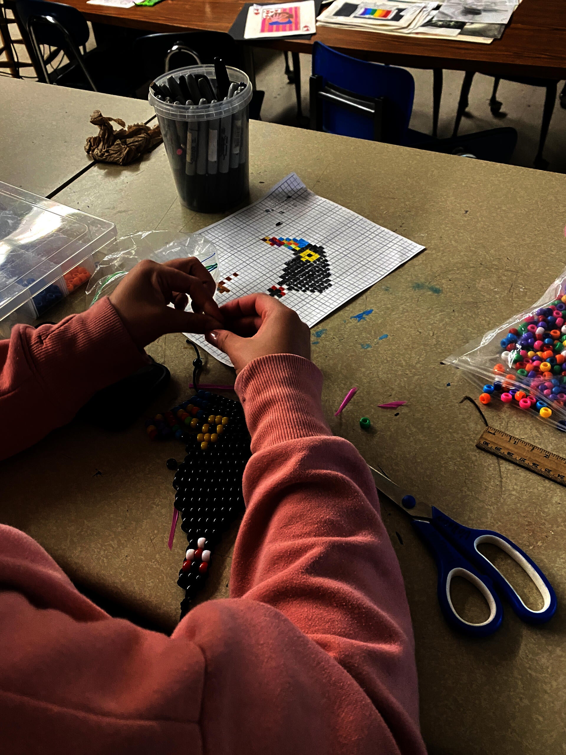 A student's hands are seen working on a pony bead toucan. A drawn design of a toucan on graph paper is seen on the desk as well. Also present are a variety of beads and markers.