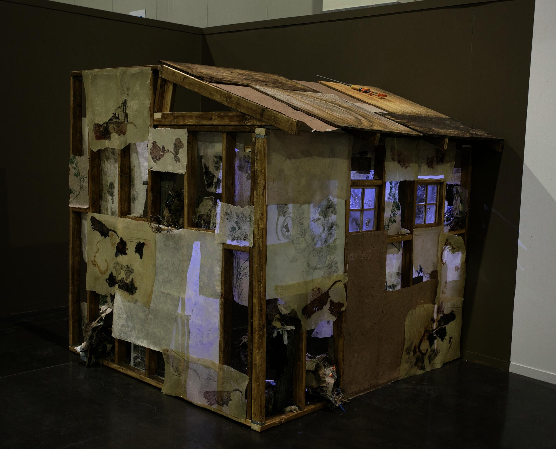 a house-like structue roughly under construction, prints of plants and roadkill line the wall, with a glow comingthrough the windows. The roof is made out of old wood and paper.