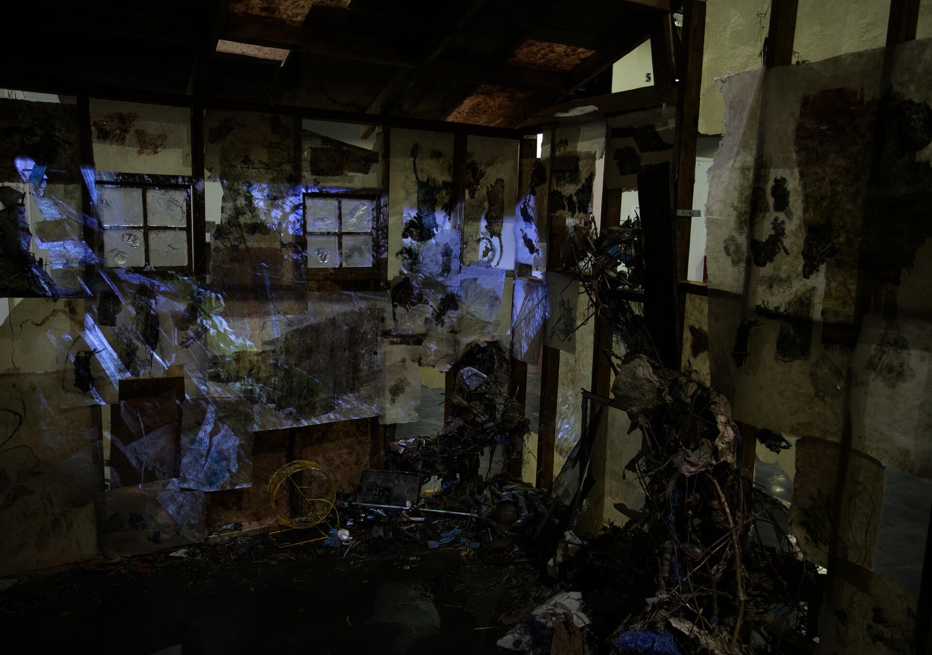 Inside a structure with exposed framing, two windows and print-lined walls, with nest-like forms going into the walls, and a projection of a pile of a pile of wood in a garden