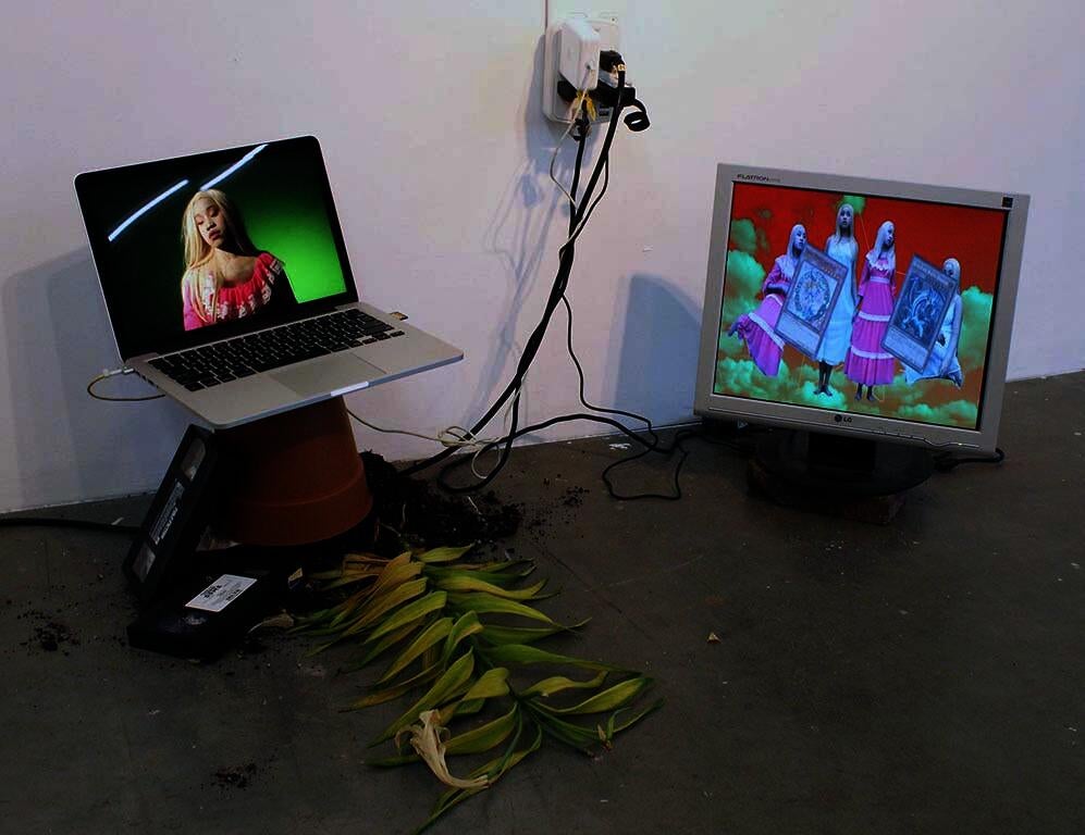 Two screens sit on the floor of the gallery. DARKMAGICIANGIRL appears on a screen to the right and Untitled (Pink Portrait) appears to the left, sitting on top of a terracotta flower pot. There are two VHS tapes to the left of the pot, and a peace-lily with potting soil spilling out from underneath it.