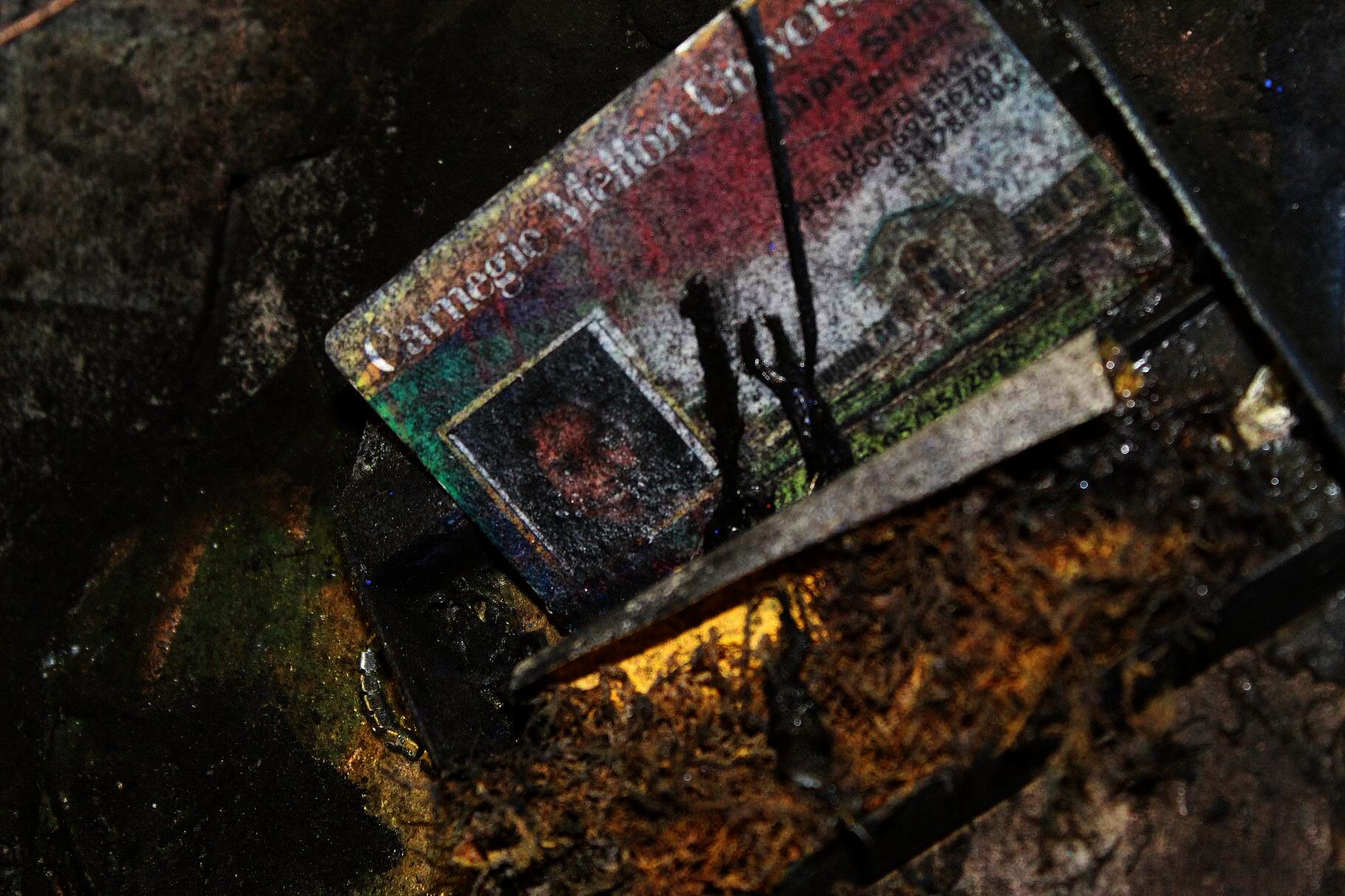 The image shows a close-up of STREETTRASH, featuring the artist's undergraduate student ID covered in thick black liquid and flecks of aerosol paint.