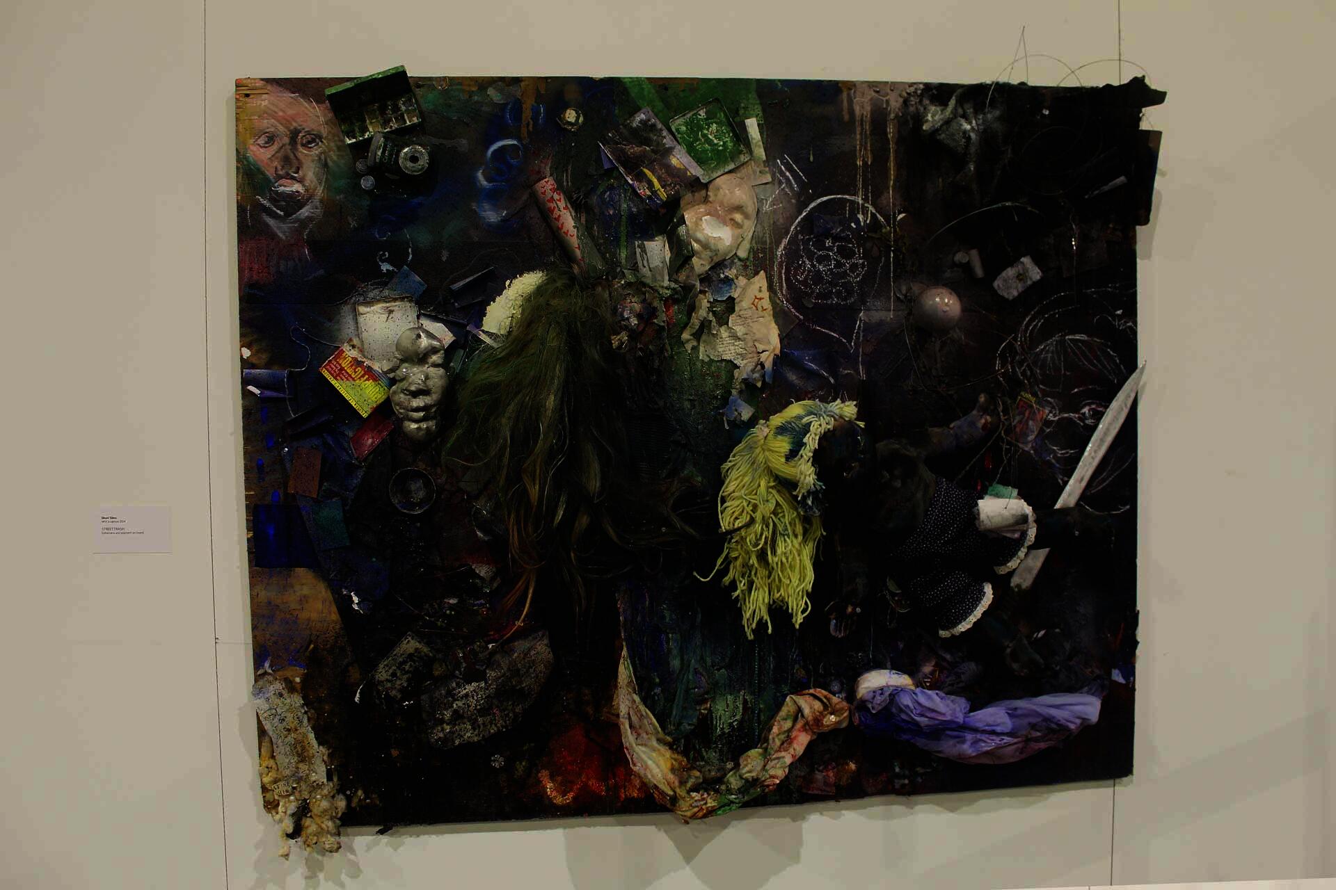 The image depicts a large painting, with dark pigment around the edges and more greenish and blueish shades of pigment towards the middle. To the center-right and upper-left of the painting, there are drawings of faces, and many objects and ephemera attached to the board as well, including a sword, cassette tapes and cases, a small digital camera, a glass orb, a small watercolor compact, and a fabric doll with blond yarn hair.