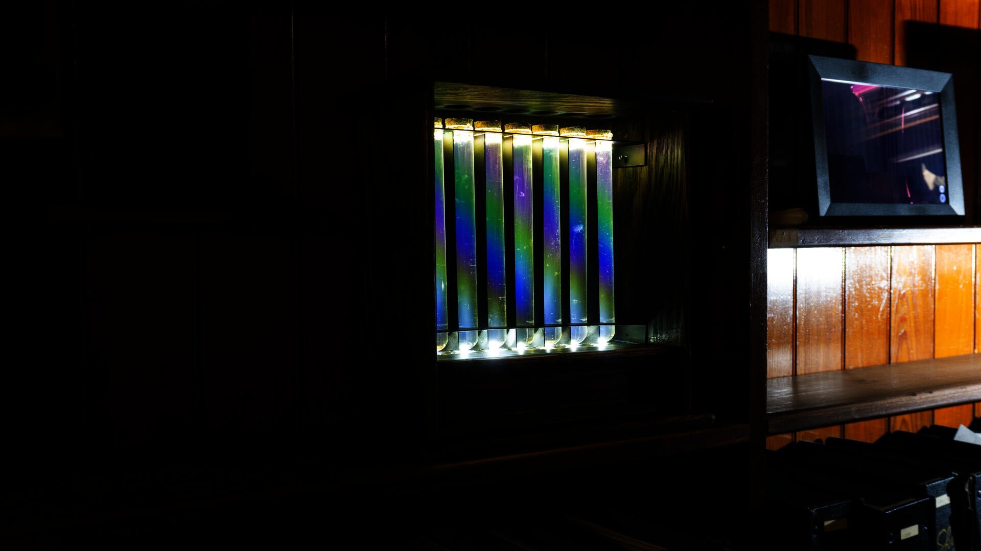 An antique-like wooden box holds a line of test tubes filled with liquids, with iridescent colors forming an arc from the device's arrangement.
