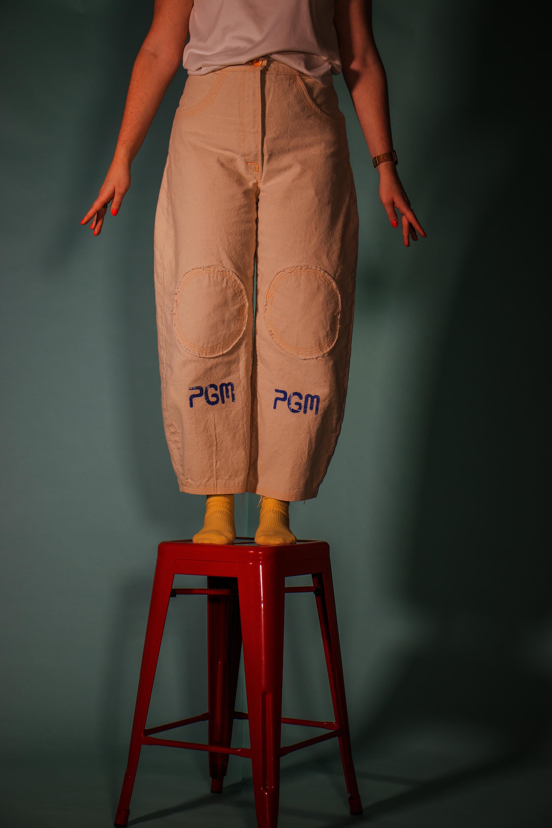 model standing on a red stool, showcasing canvas pants with her arms at her sides. you can't see her face, but you can see her yellow socks and the blue block print of the logo "PGM" under big circular knee pads finished with an orange button.