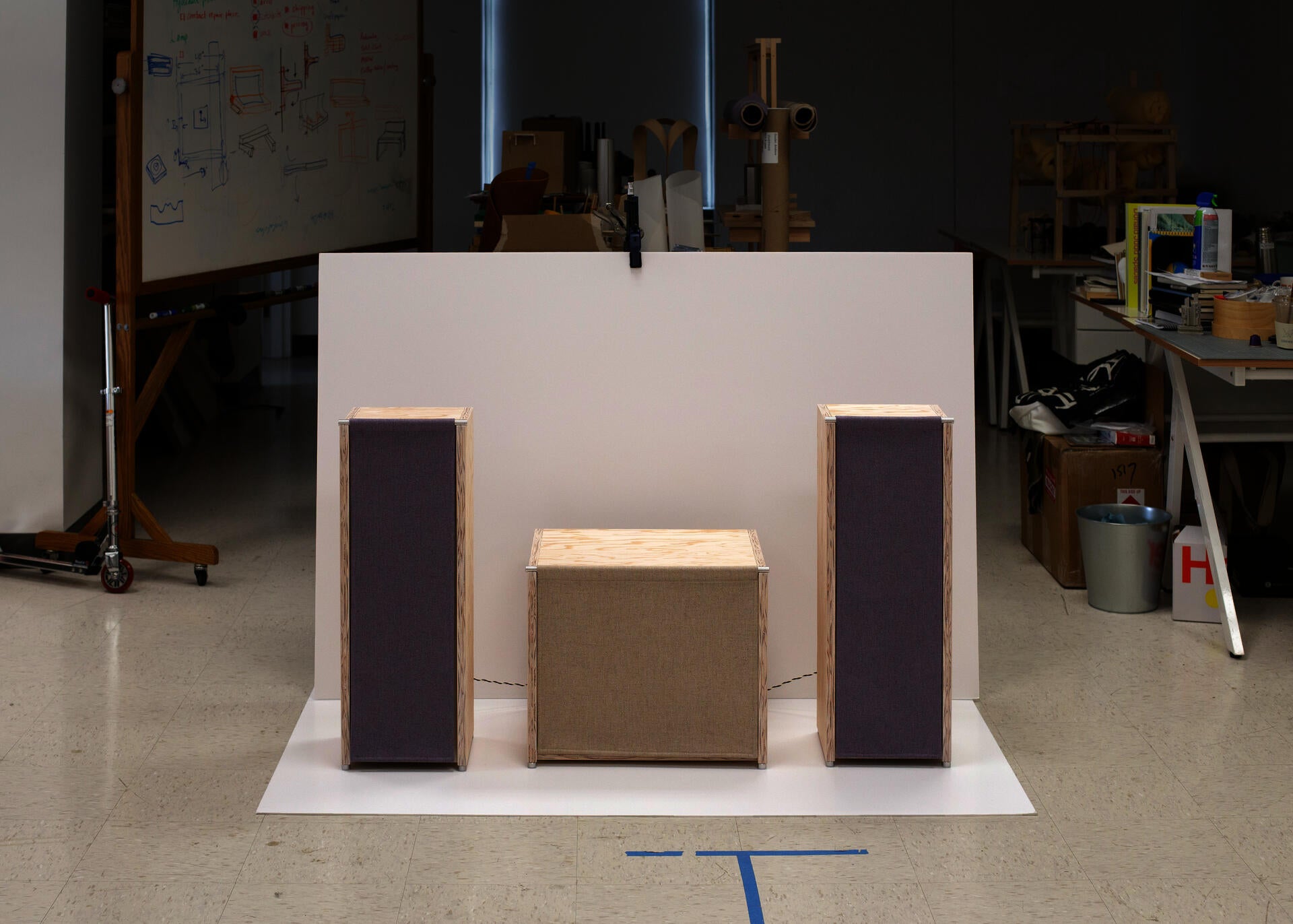 Two pale plywood speakers with hanging fabric fronts symmetrically flank a lower cabinet with a similar fabric front in a design studio, set against a large white backdrop with sketches, tools, and prototype furniture visible in the background.