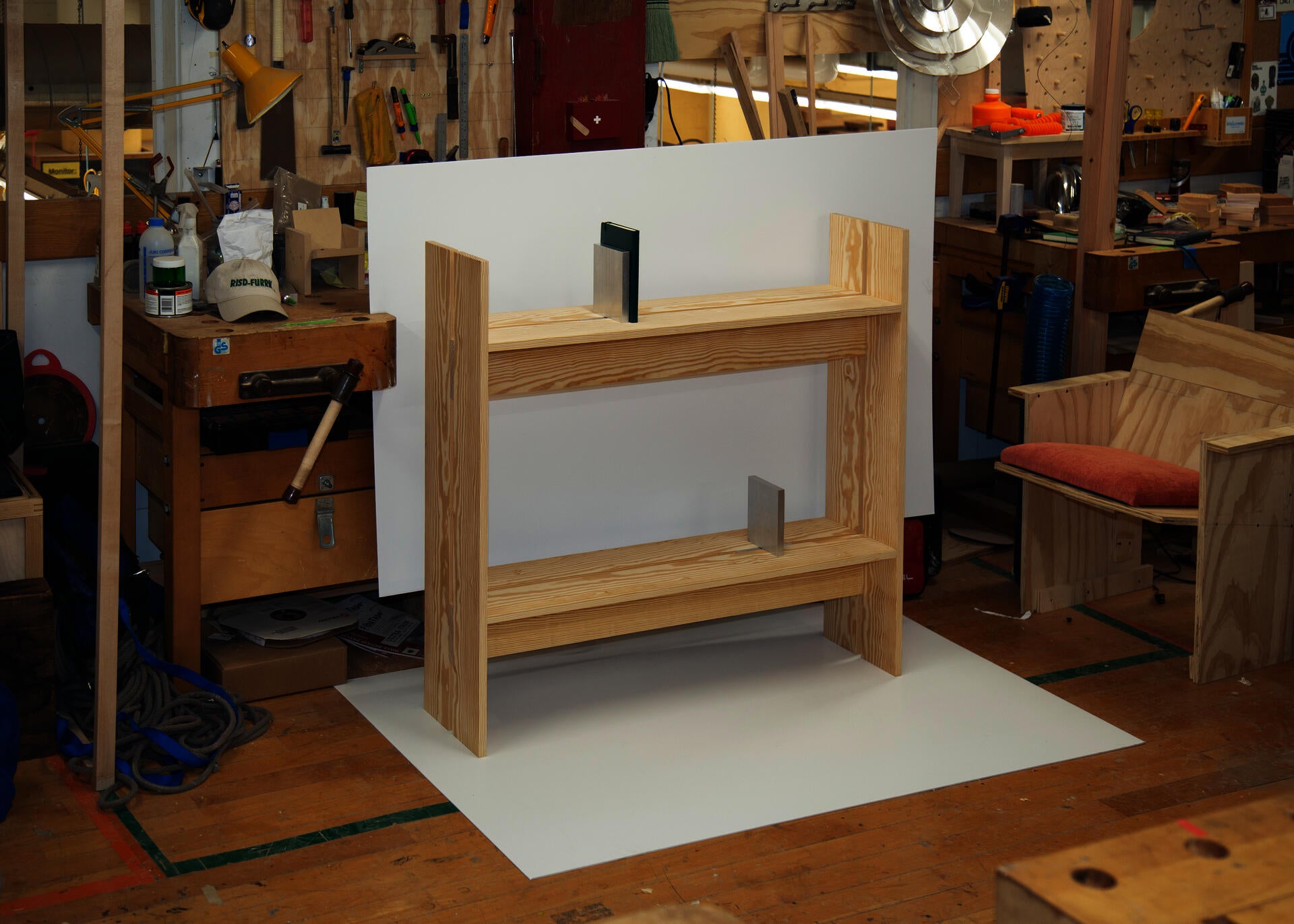 A pale wood shelving unit with integrated vertical book ends stands in a woodworking studio, displayed against a white backdrop amid workbenches, tools, and partially finished furniture prototypes.